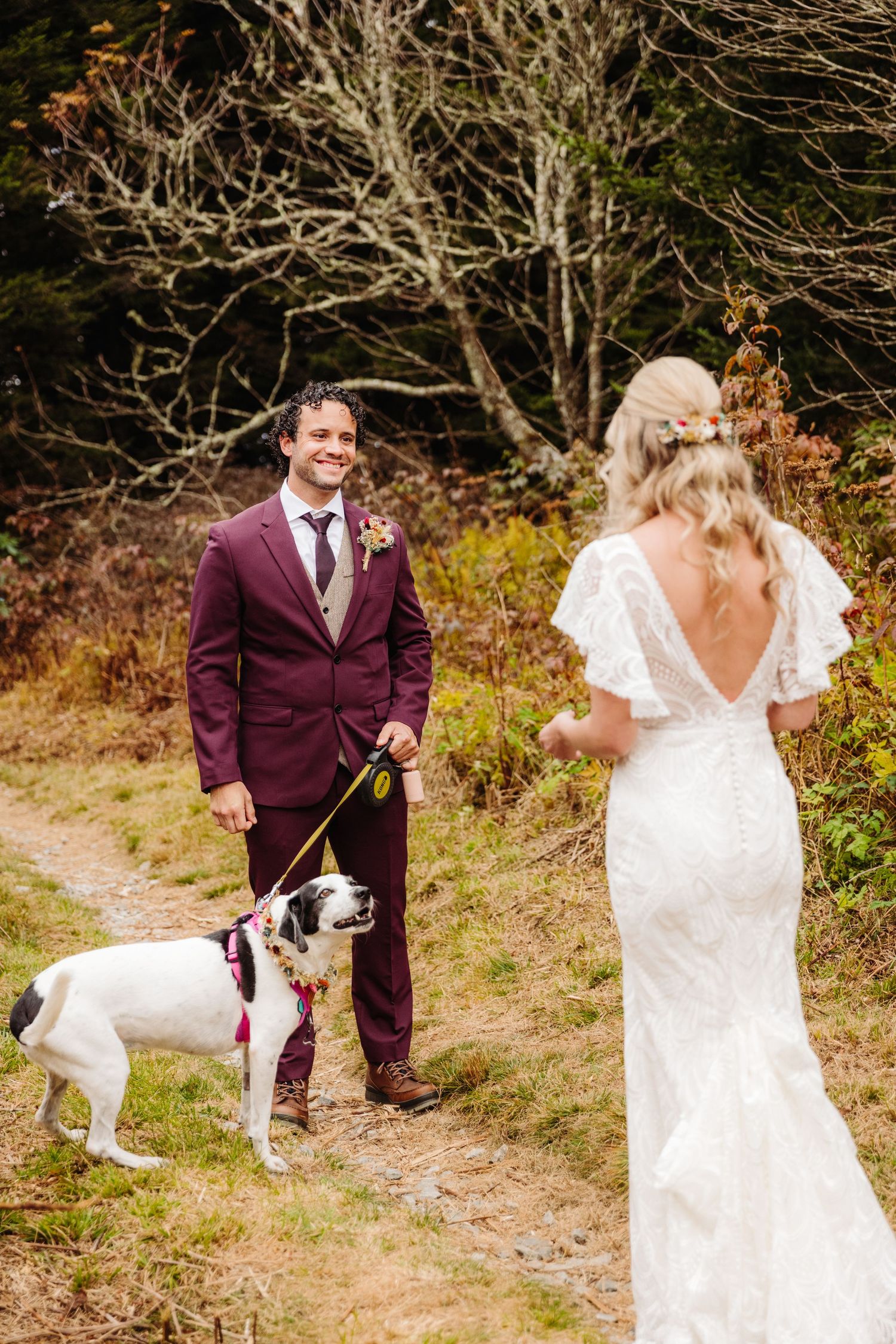 Wedding couple and their dog pose on rustic trail surrounded by autumn foliage.