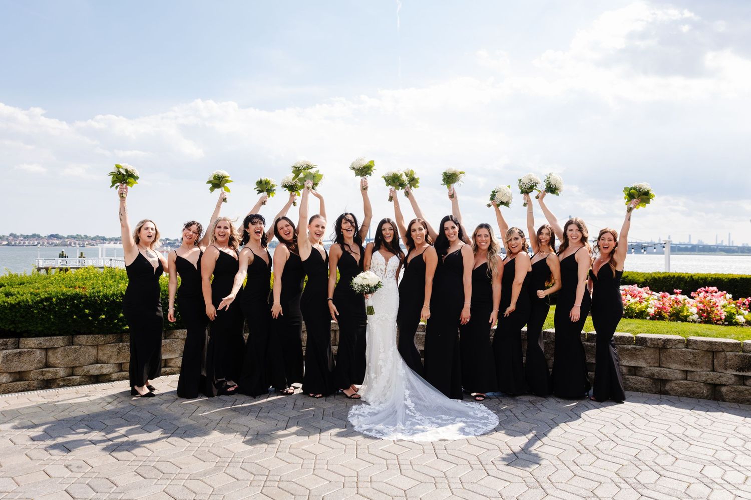Wedding party members dressed in black pose together outdoors in a line on a sunny day.