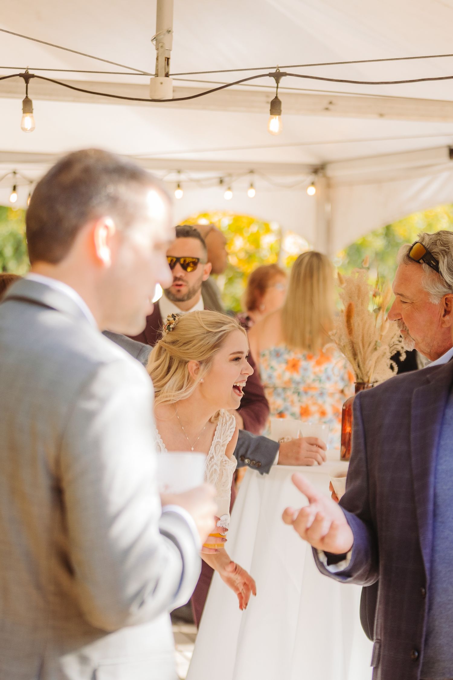 Guests mingling under a white tent with string lights at an outdoor reception.