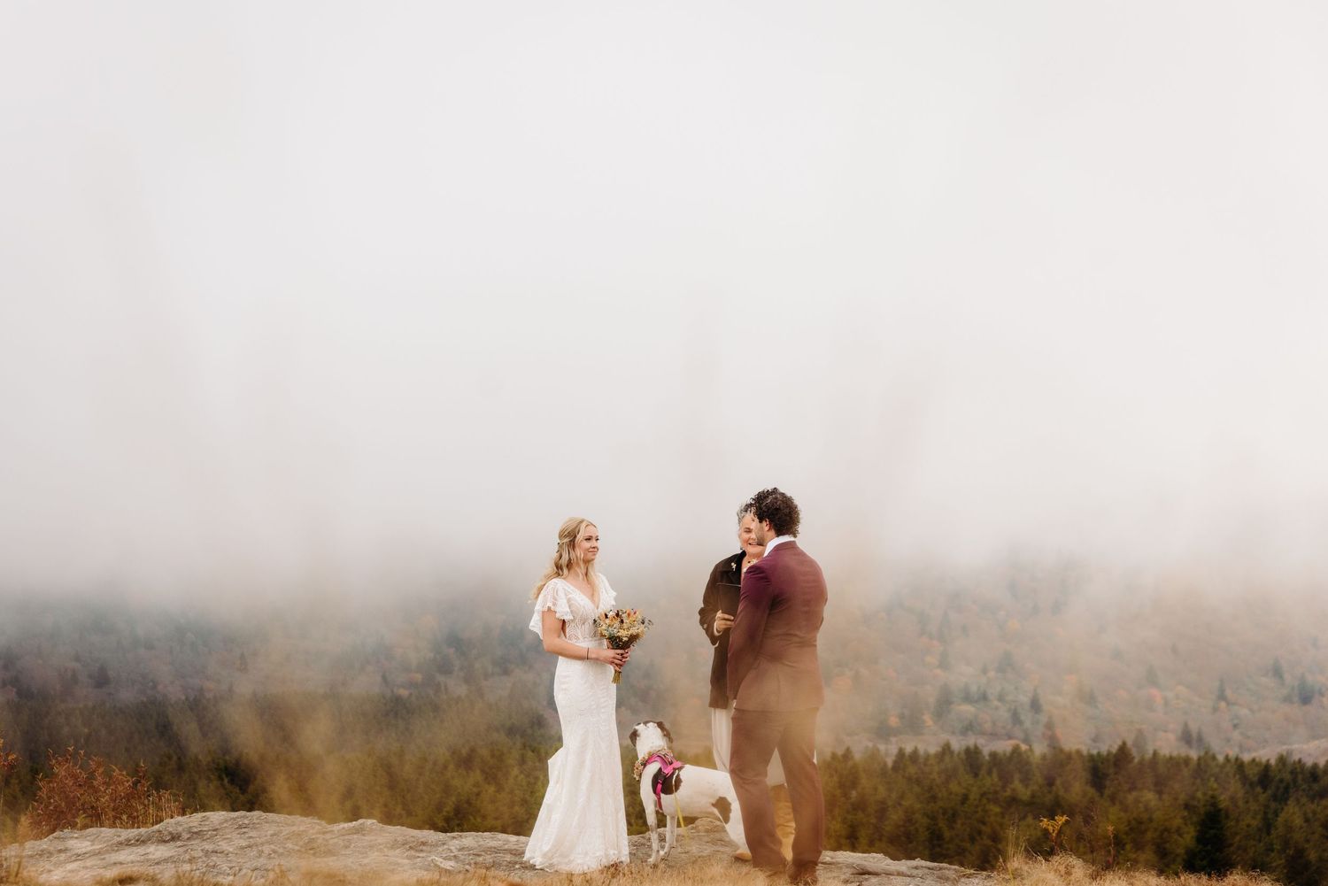 Misty mountain wedding ceremony with couple and dog against foggy backdrop.