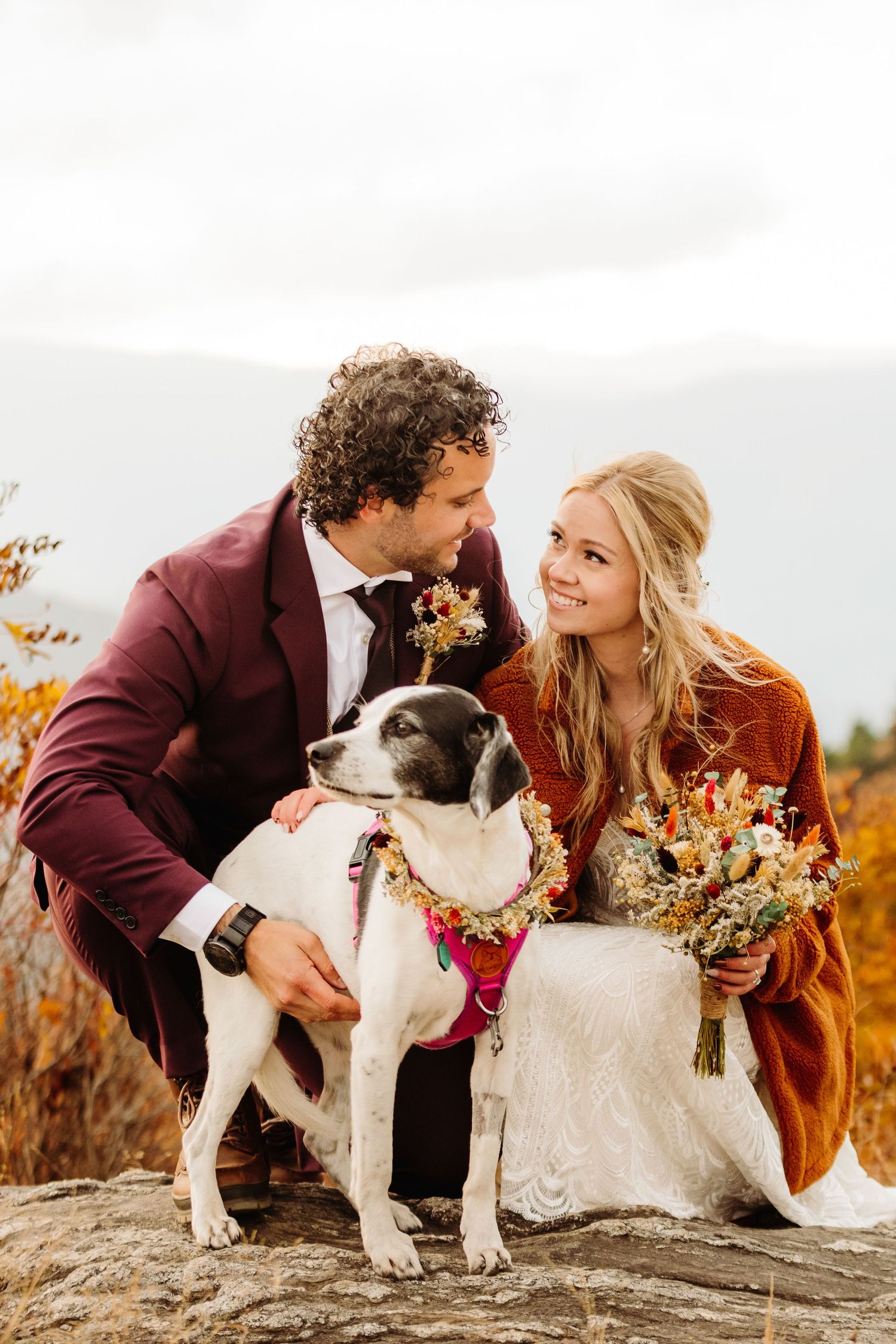Couple poses with their dog wearing floral collar on mountain summit in autumn.