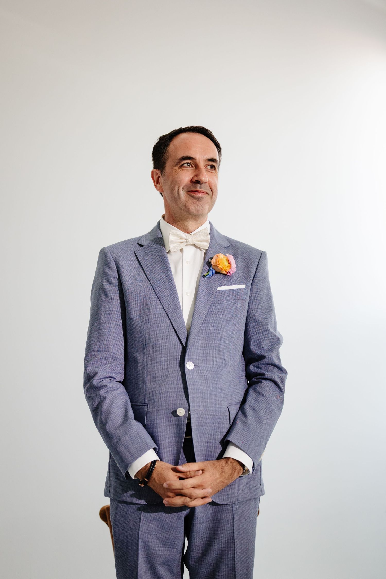 Groom in a light grey suit and white shirt smiles while standing against a white backdrop.