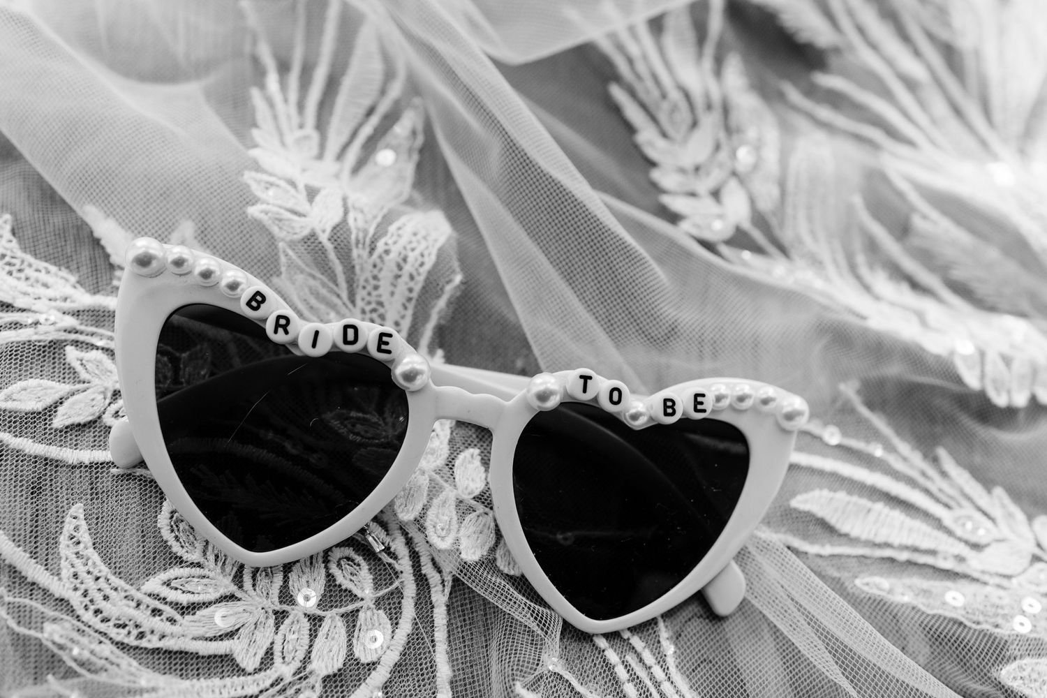 Heart-shaped white bride sunglasses resting on delicate lace fabric.