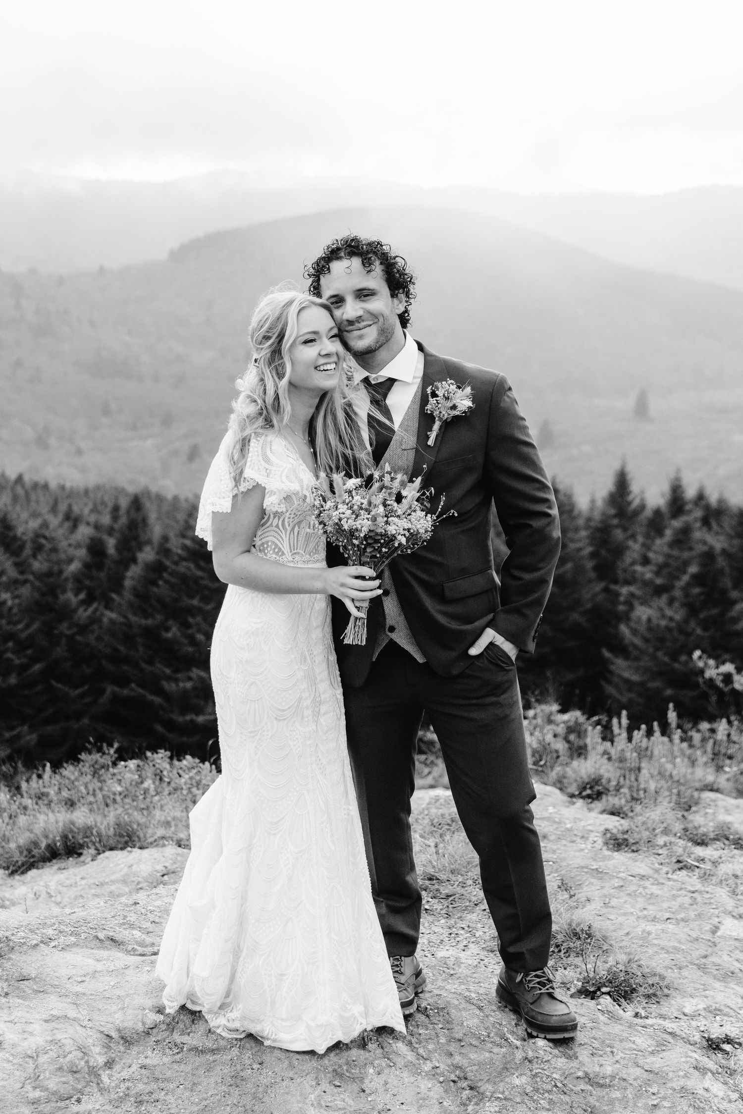 Black and white portrait of couple on mountain summit with evergreen forest backdrop.