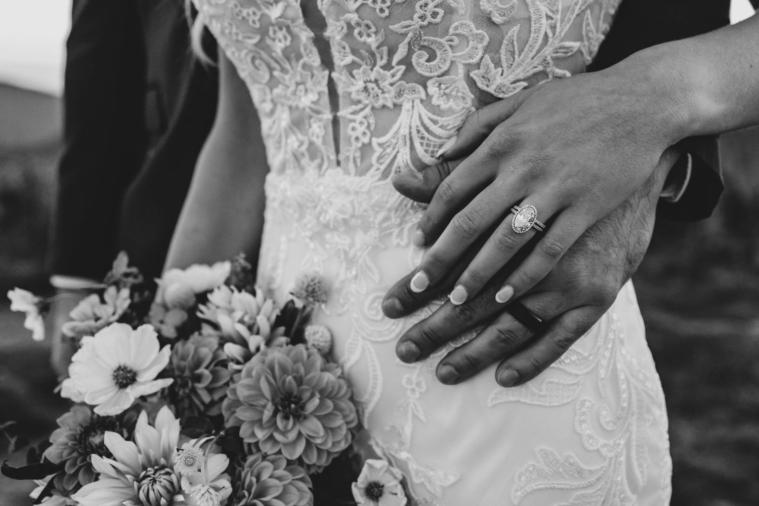 Black and white detail of hands holding a bouquet against a lace wedding dress.