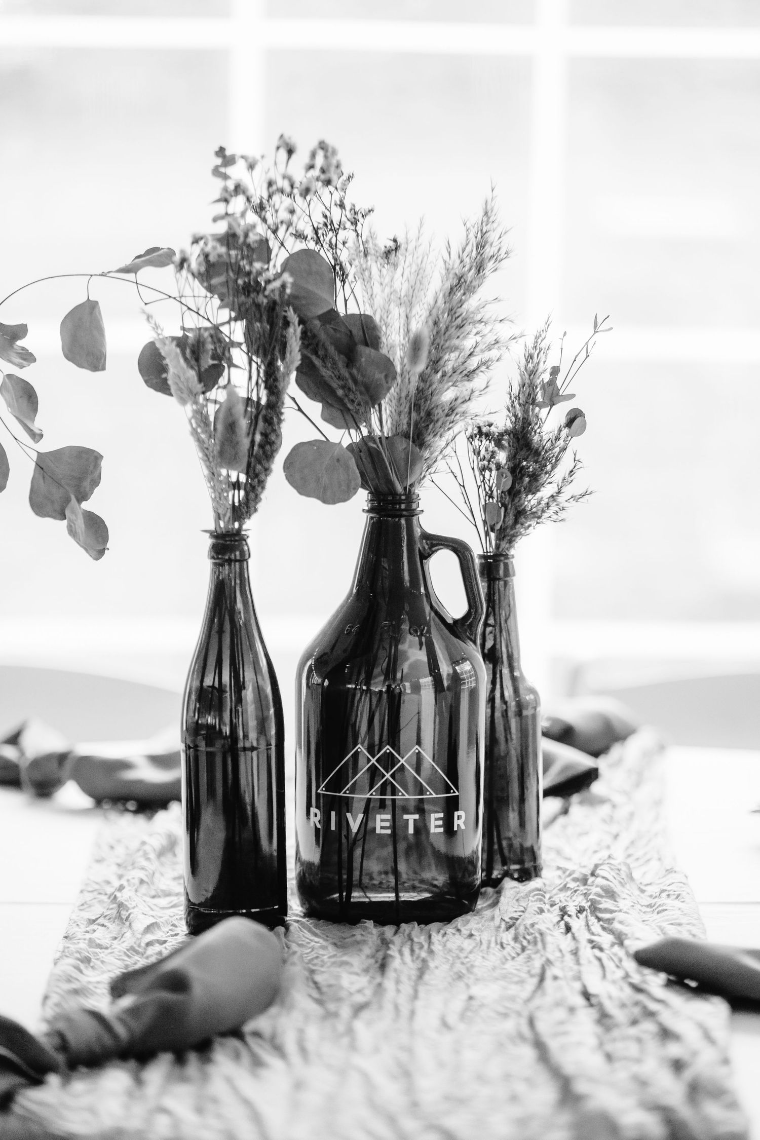 Black and white image of decorative bottles with dried flowers and greenery arranged on a textured surface.