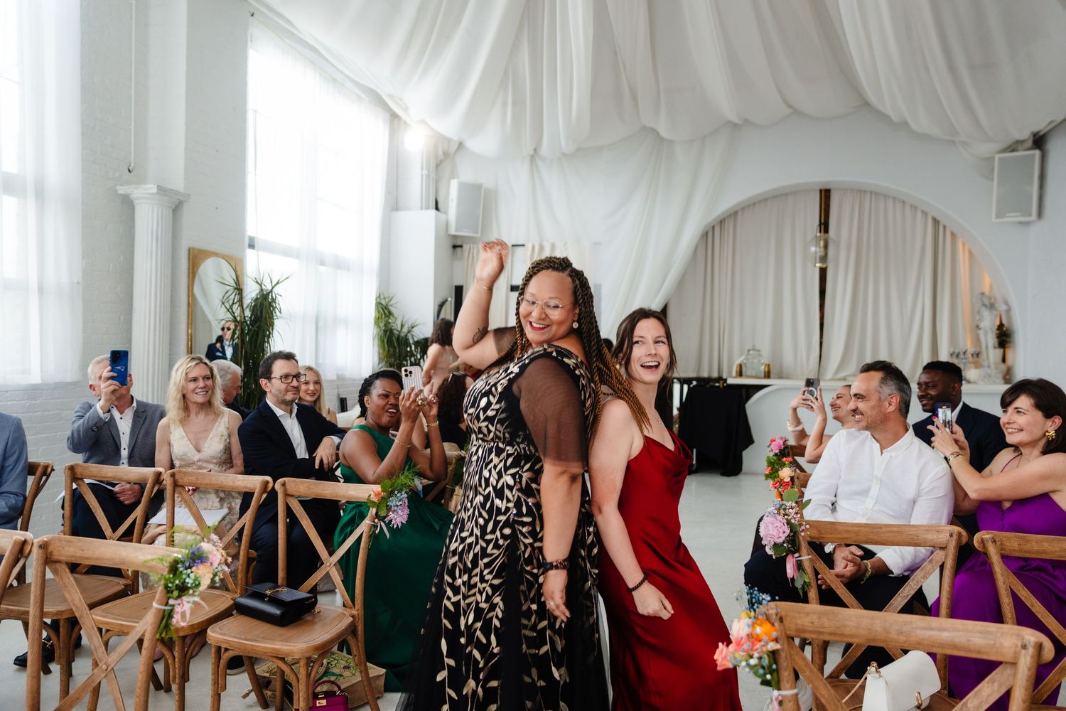 Wedding reception guests dance and celebrate in a white-draped venue with rustic wooden chairs.
