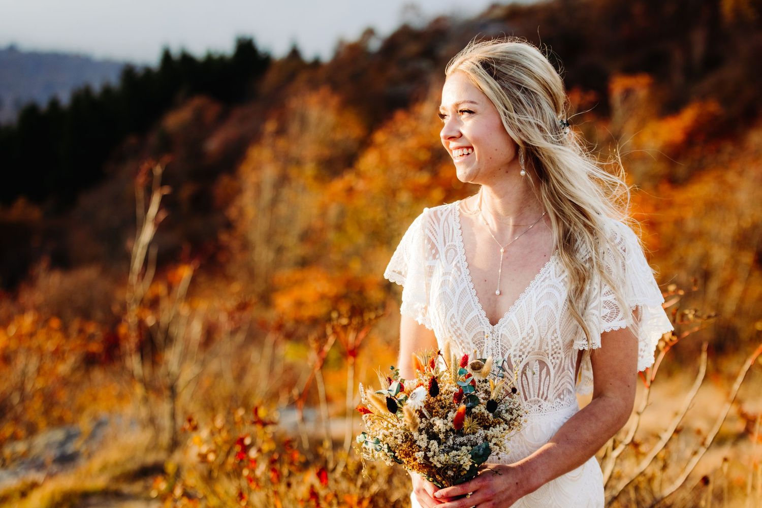 Bride in lace dress poses in golden autumn mountain field at sunset.
