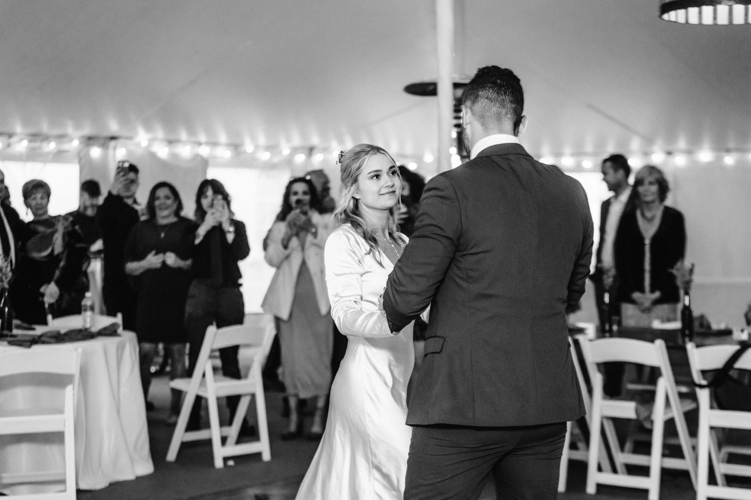 Couple shares their first dance at wedding reception under tent with guests watching.