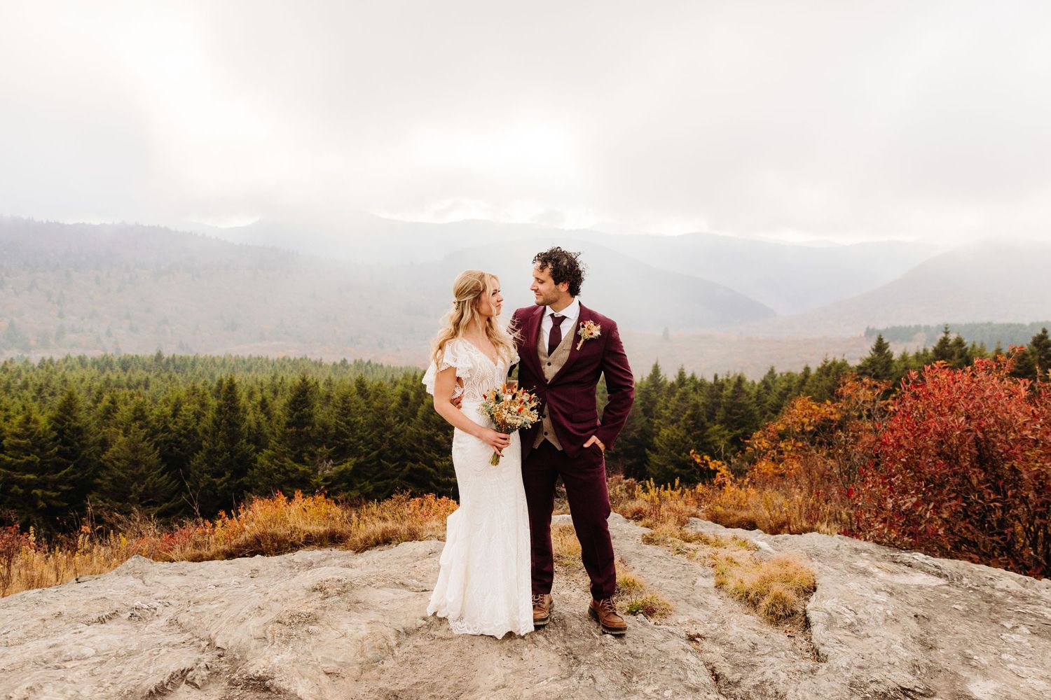 Wedding couple stands on rocky overlook with dogs surrounded by autumn mountain scenery.
