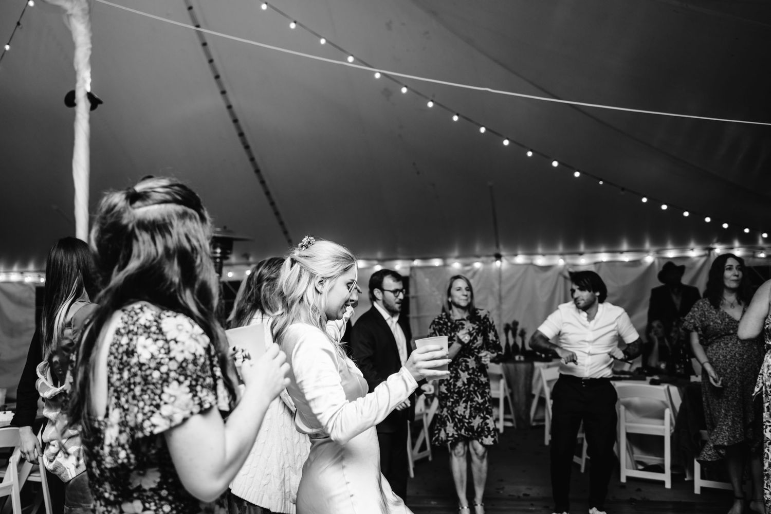 Wedding guests dance and celebrate under a tent with string lights at an evening reception.