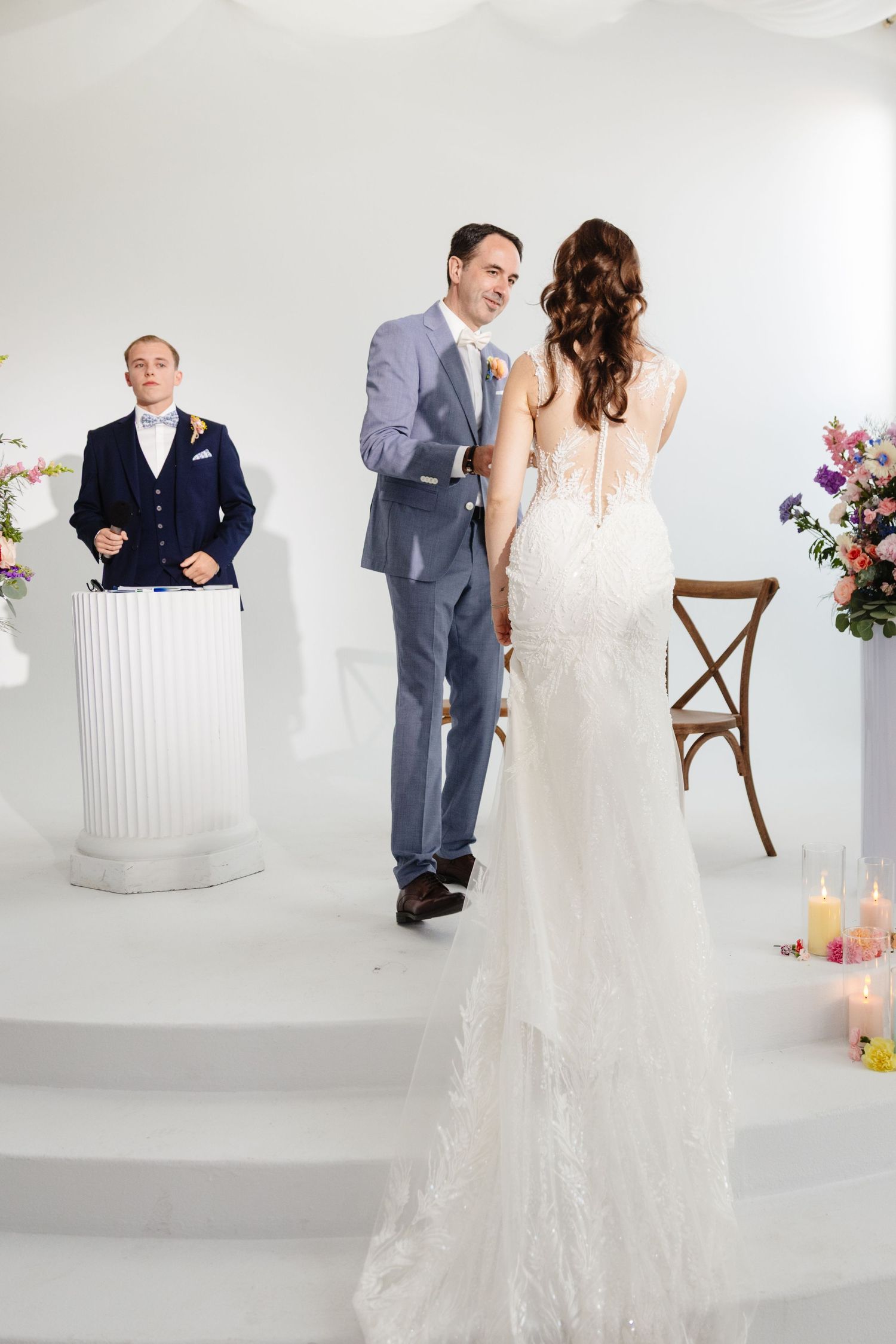 Couple in wedding attire poses in an elegant white studio setting with floral arrangements.