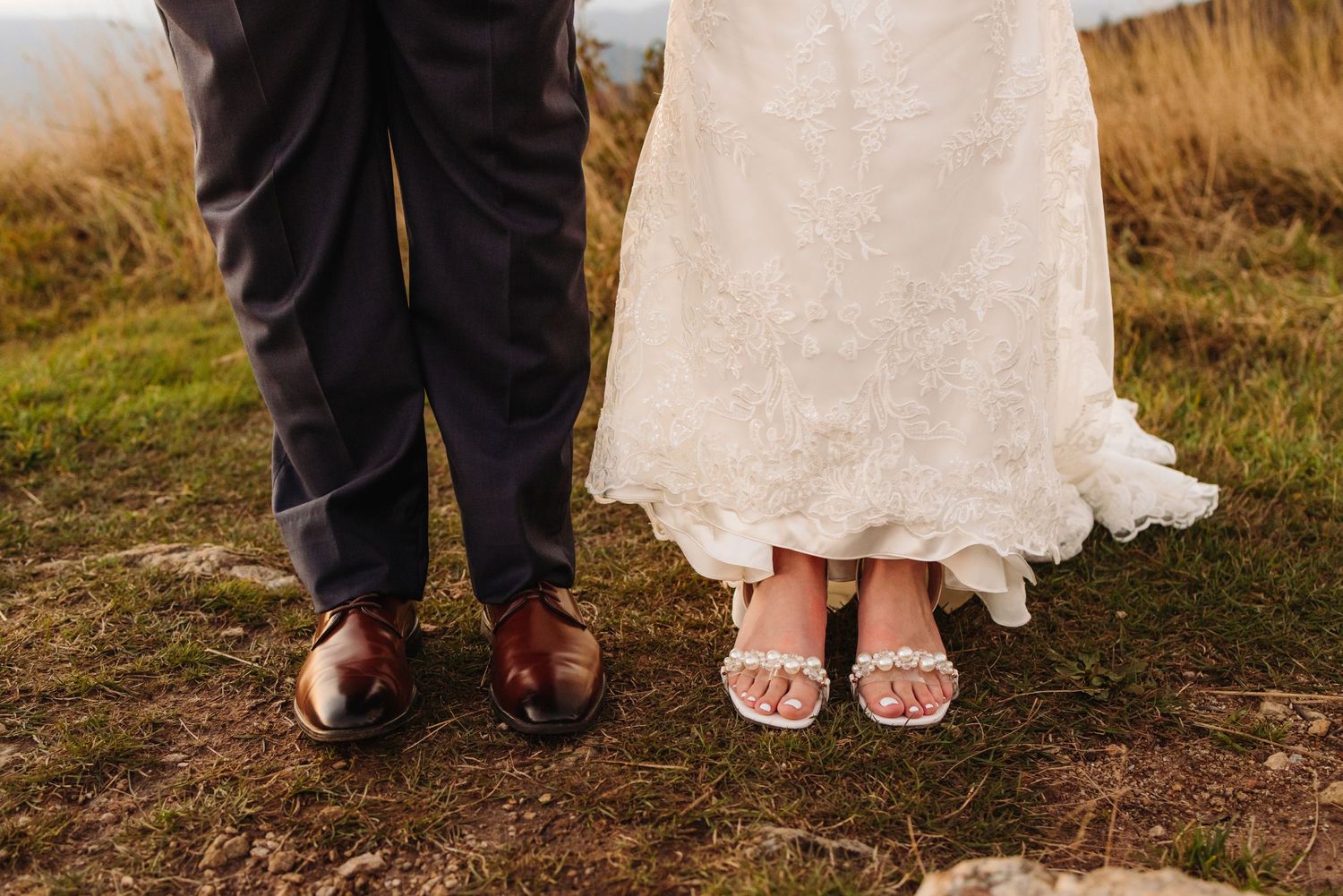 Wedding couple's feet showing formal dress shoes and sparkly bridal sandals on mountain terrain.