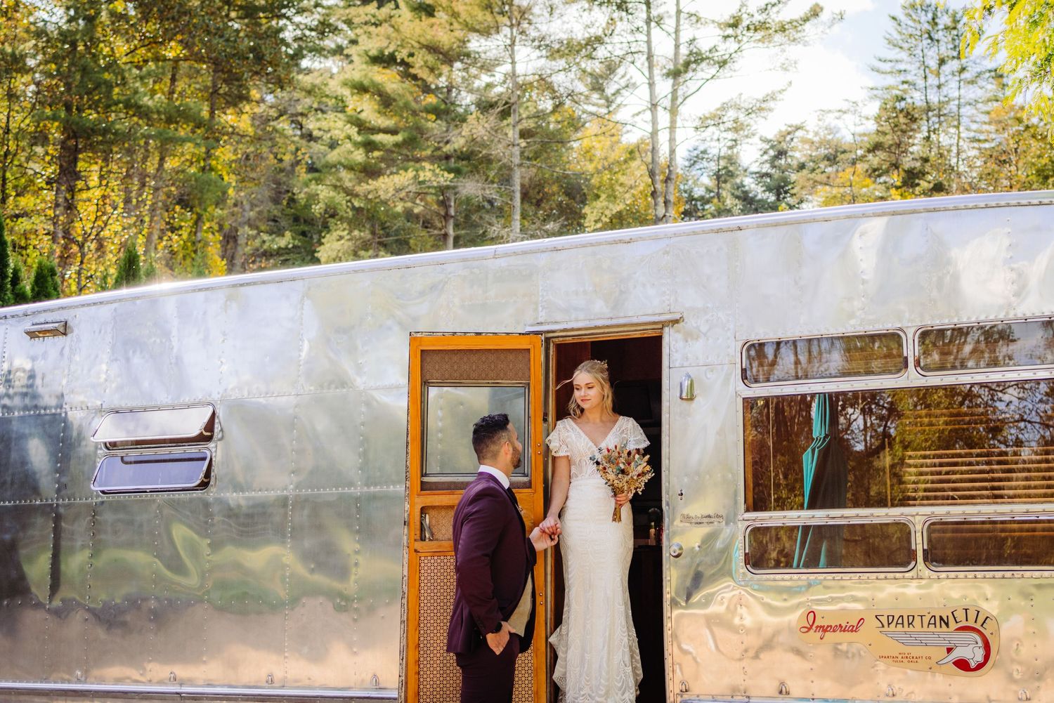 A couple poses outside a vintage silver Airstream trailer in a wooded setting.