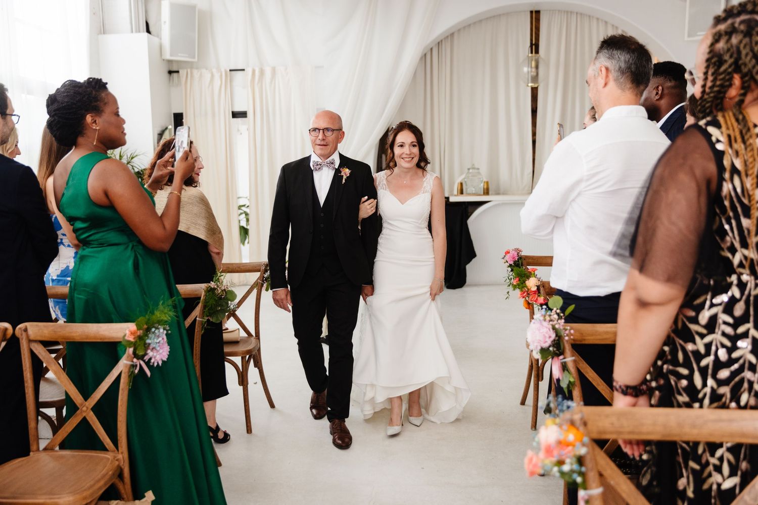 Guests gather in a white-draped venue with wooden chairs for a wedding celebration.