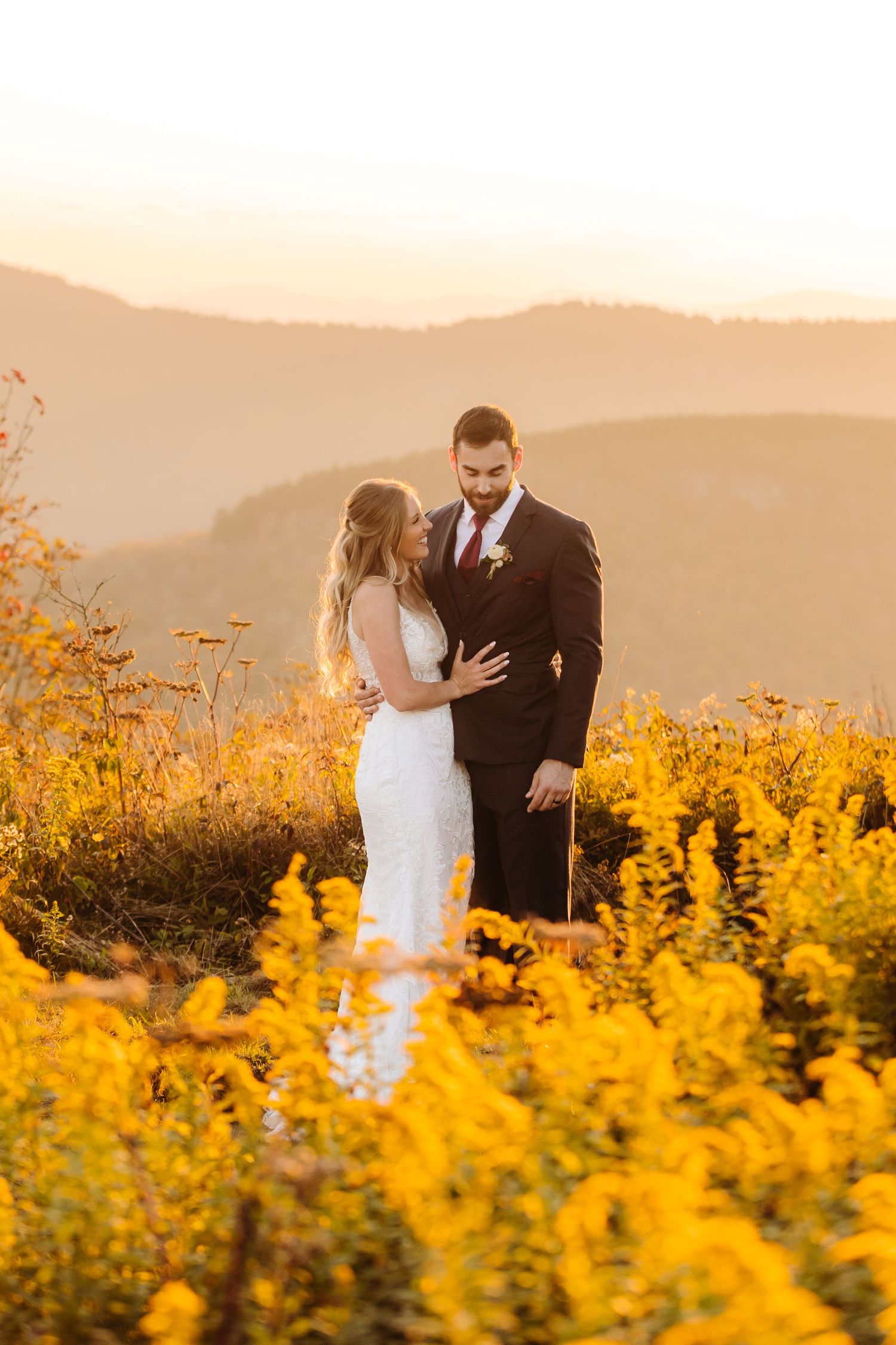 Newlyweds share intimate moment among golden wildflowers with mountain vista backdrop.