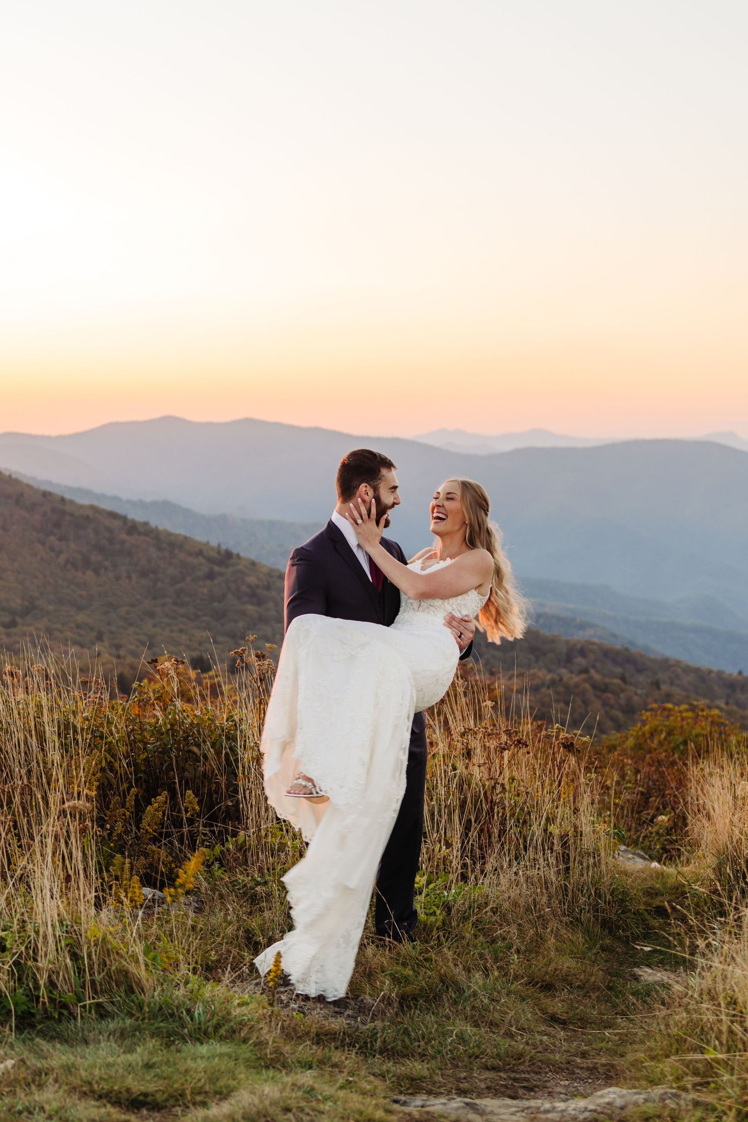 A romantic wedding couple embraces against a backdrop of misty mountain ranges at sunset.