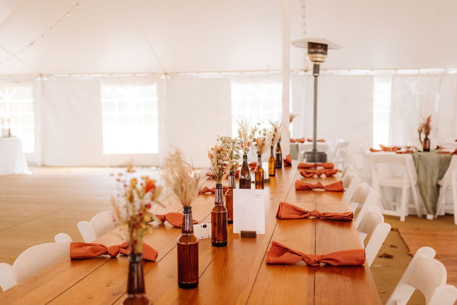 Wedding reception tables set with amber bottles and dried flower centerpieces under a white tent.