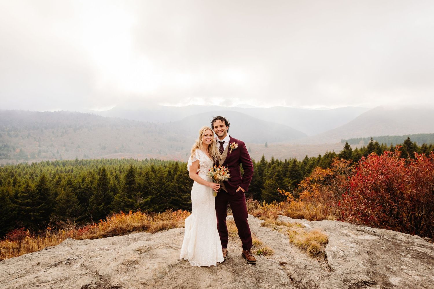 Newlyweds pose together on mountain peak with dog while surrounded by fall foliage.