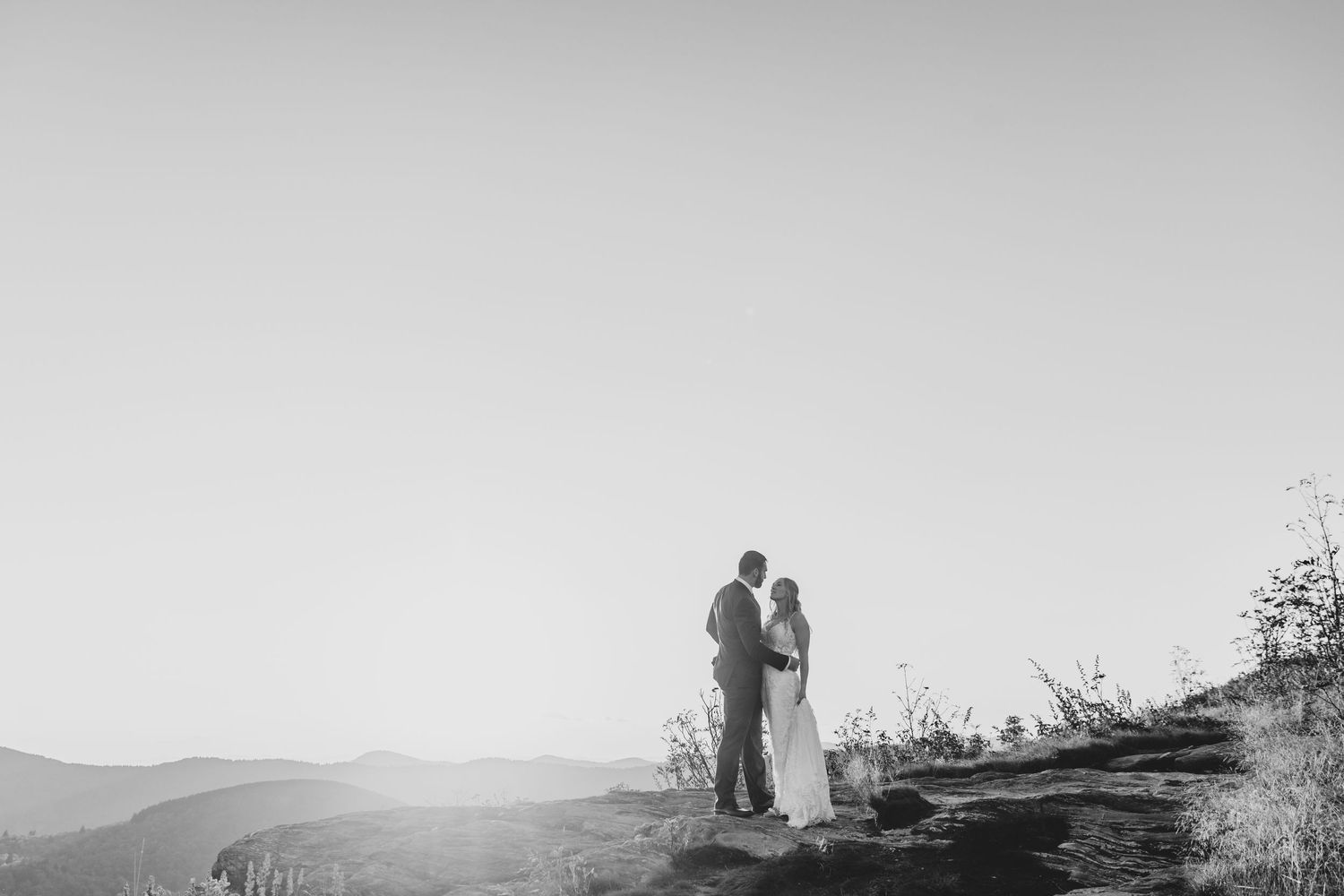 Silhouette of wedding couple standing on mountain ridge at sunset in black and white.
