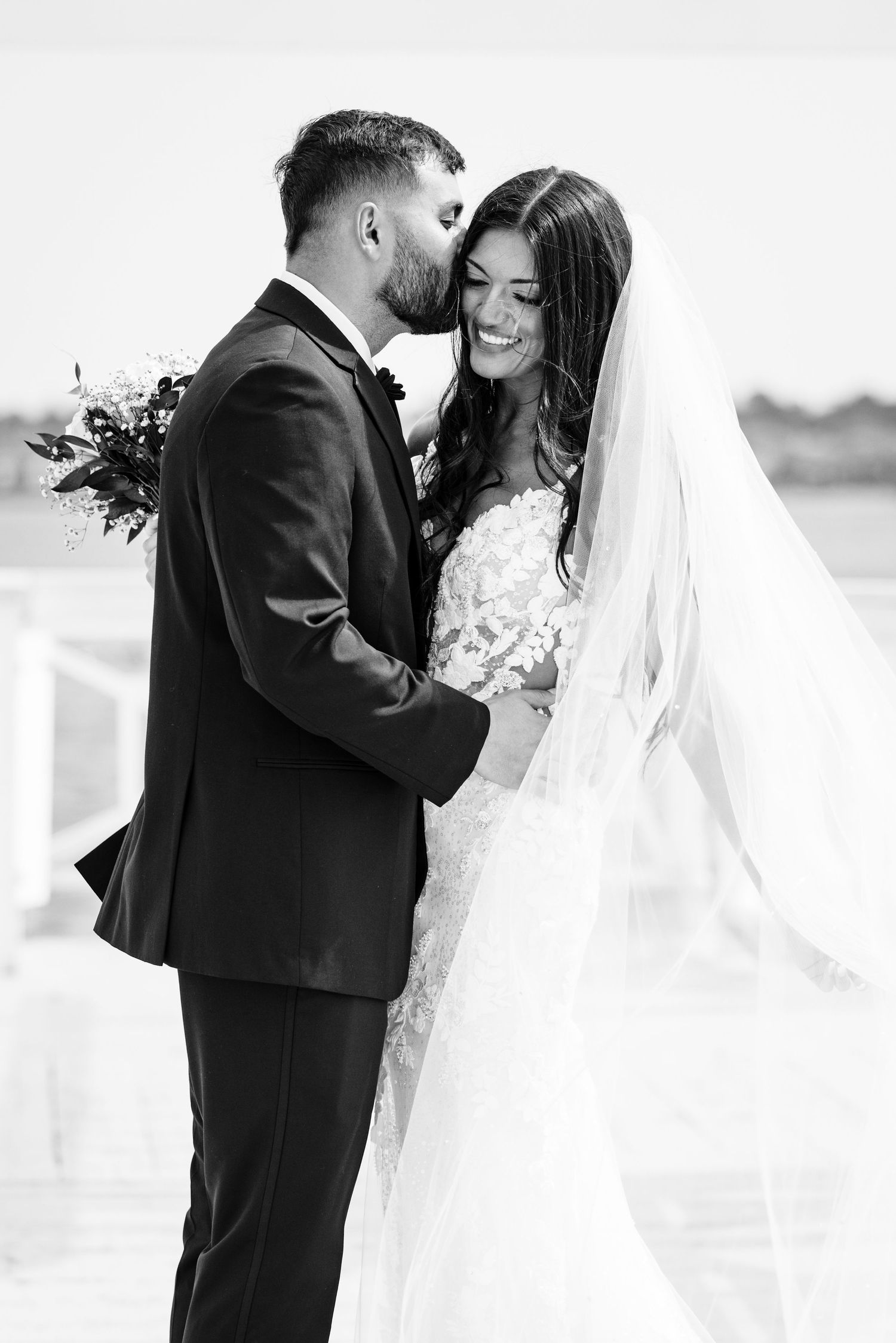 Black and white portrait of newlyweds embracing with flowing veil in the breeze.