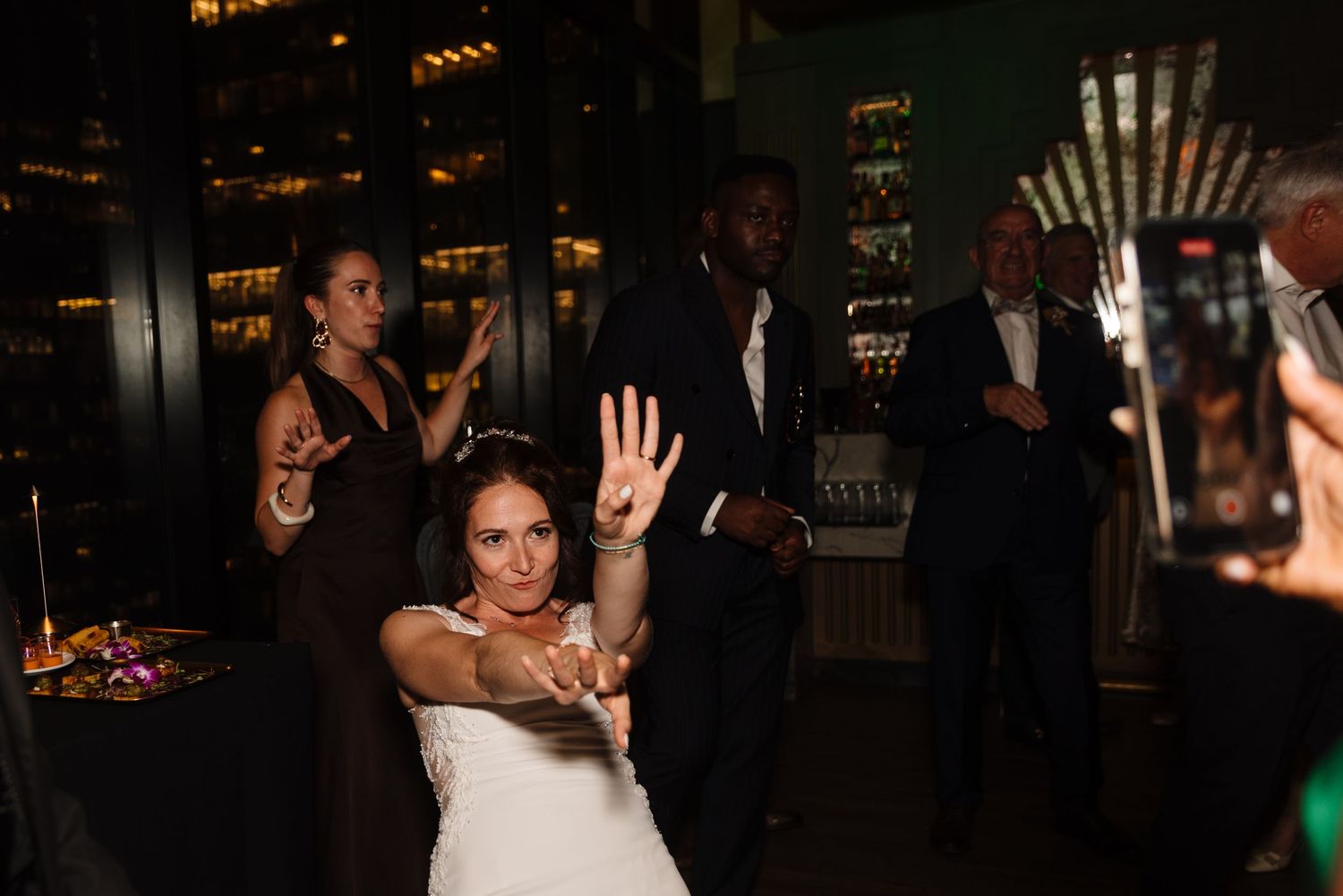 Bride and wedding guests dancing with raised arms on a dark dance floor.
