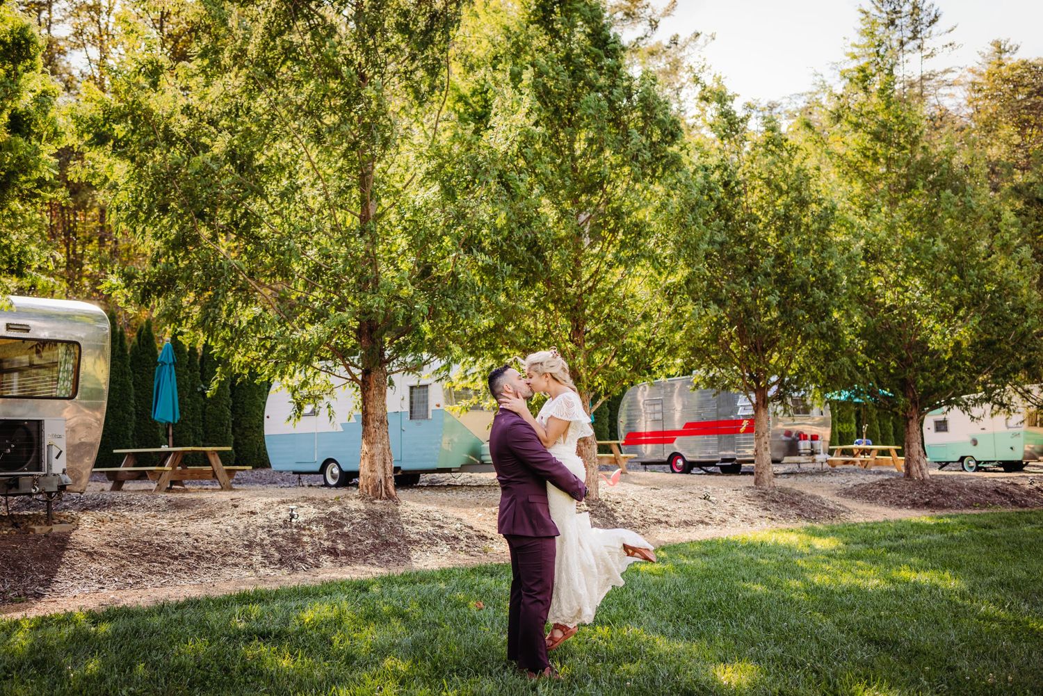 A romantic embrace in a campground setting with vintage trailers and lush trees in the background.