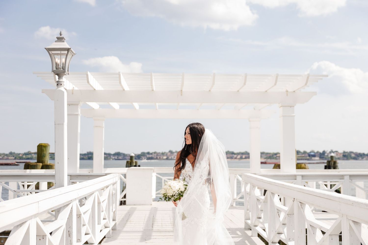 Bride in white dress holds bouquet while standing on waterfront deck under pergola.