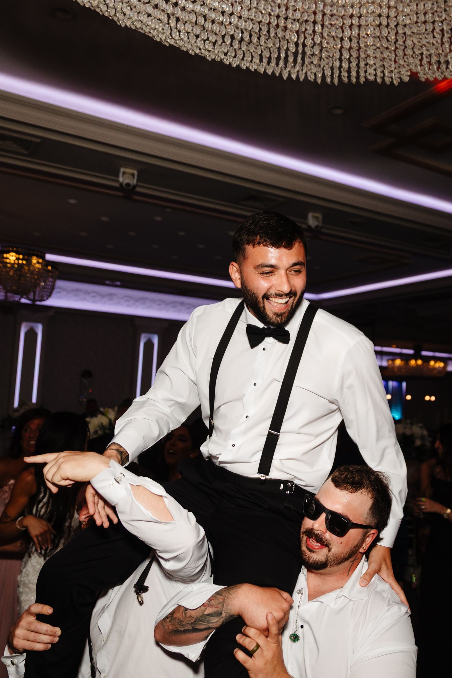 Groom in white tuxedo and suspenders being lifted by guests during wedding celebration.