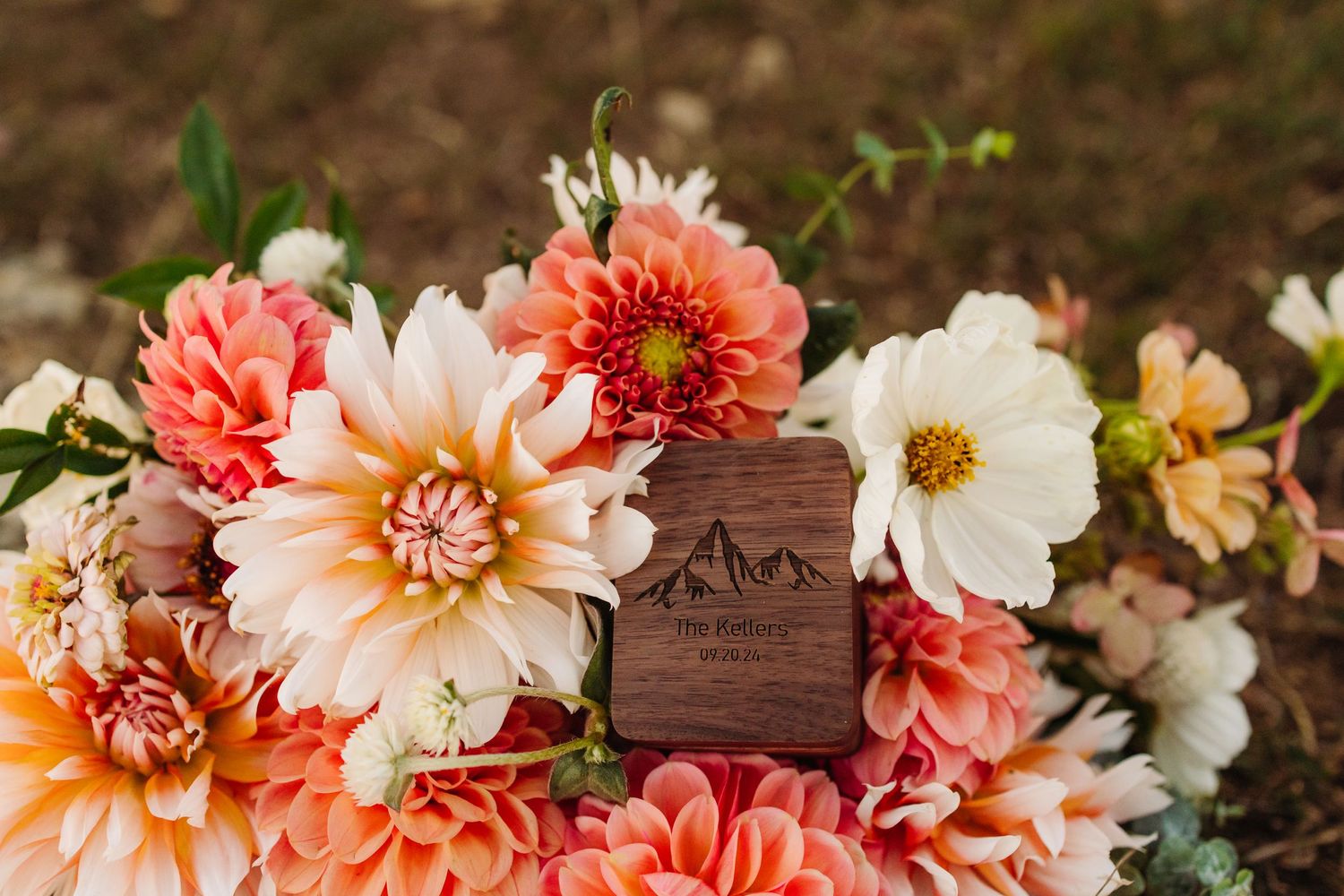 Wooden ring box surrounded by coral and peach dahlias and white cosmos flowers.