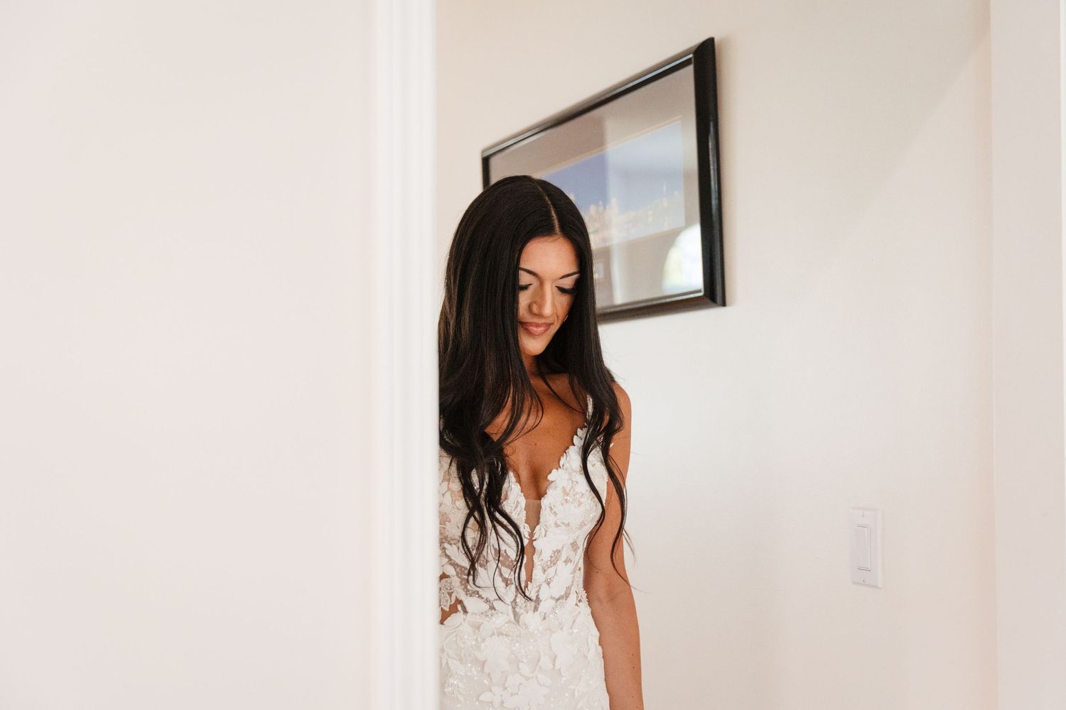 Bride in an intricate white lace dress poses near a window during wedding day preparations.