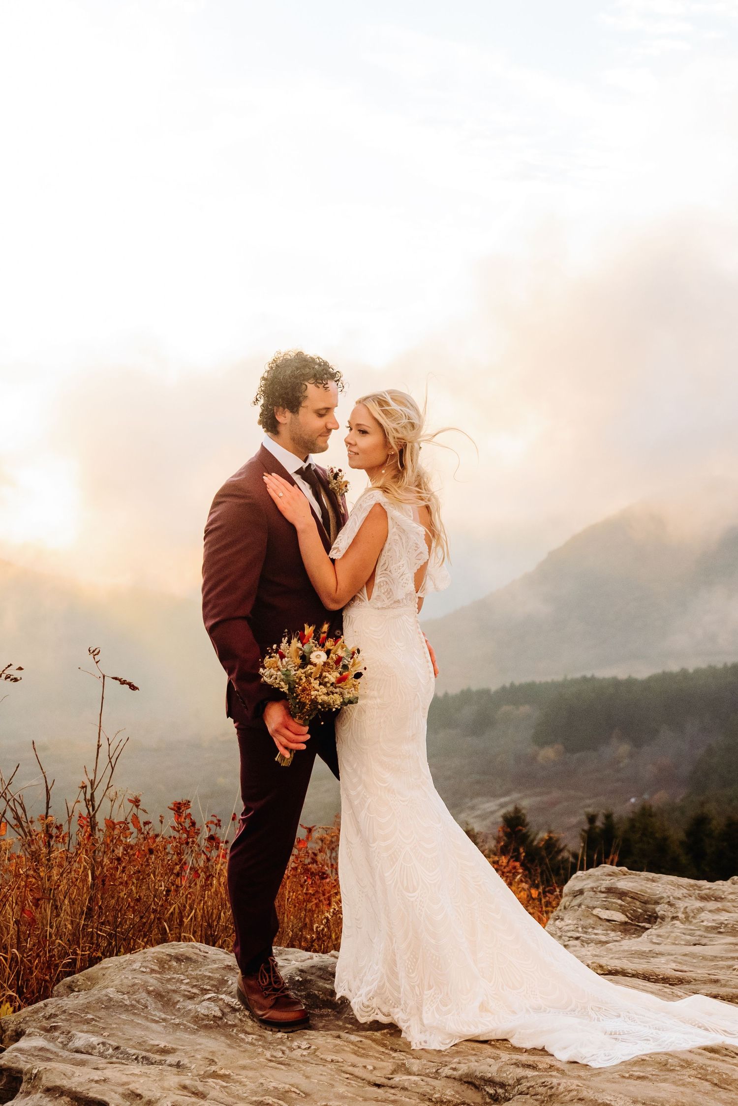 Couple embraces on mountain cliff with stunning sunset and mountain landscape behind them.