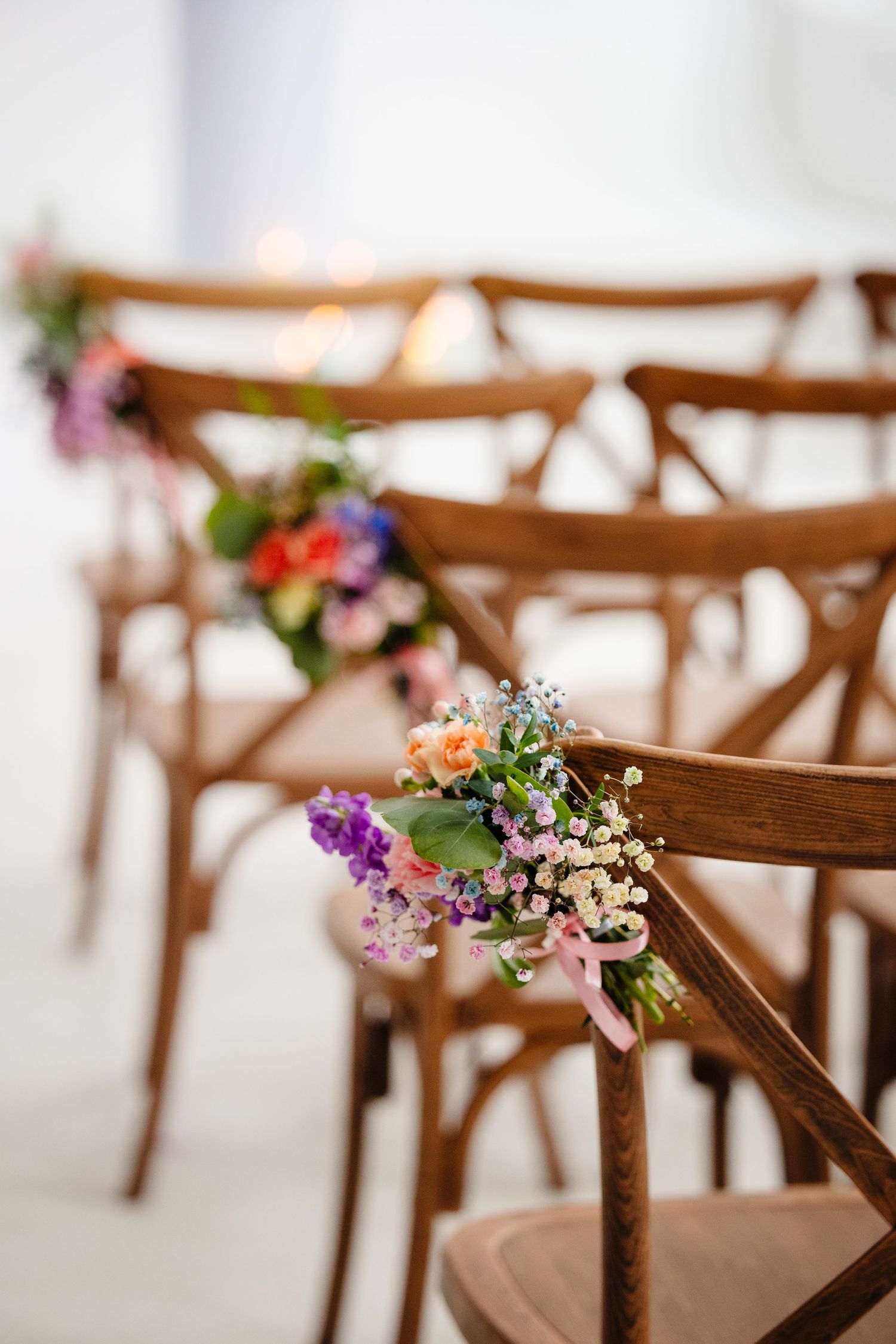 Wooden cross-back chairs decorated with colorful wildflower bouquets for a wedding ceremony.
