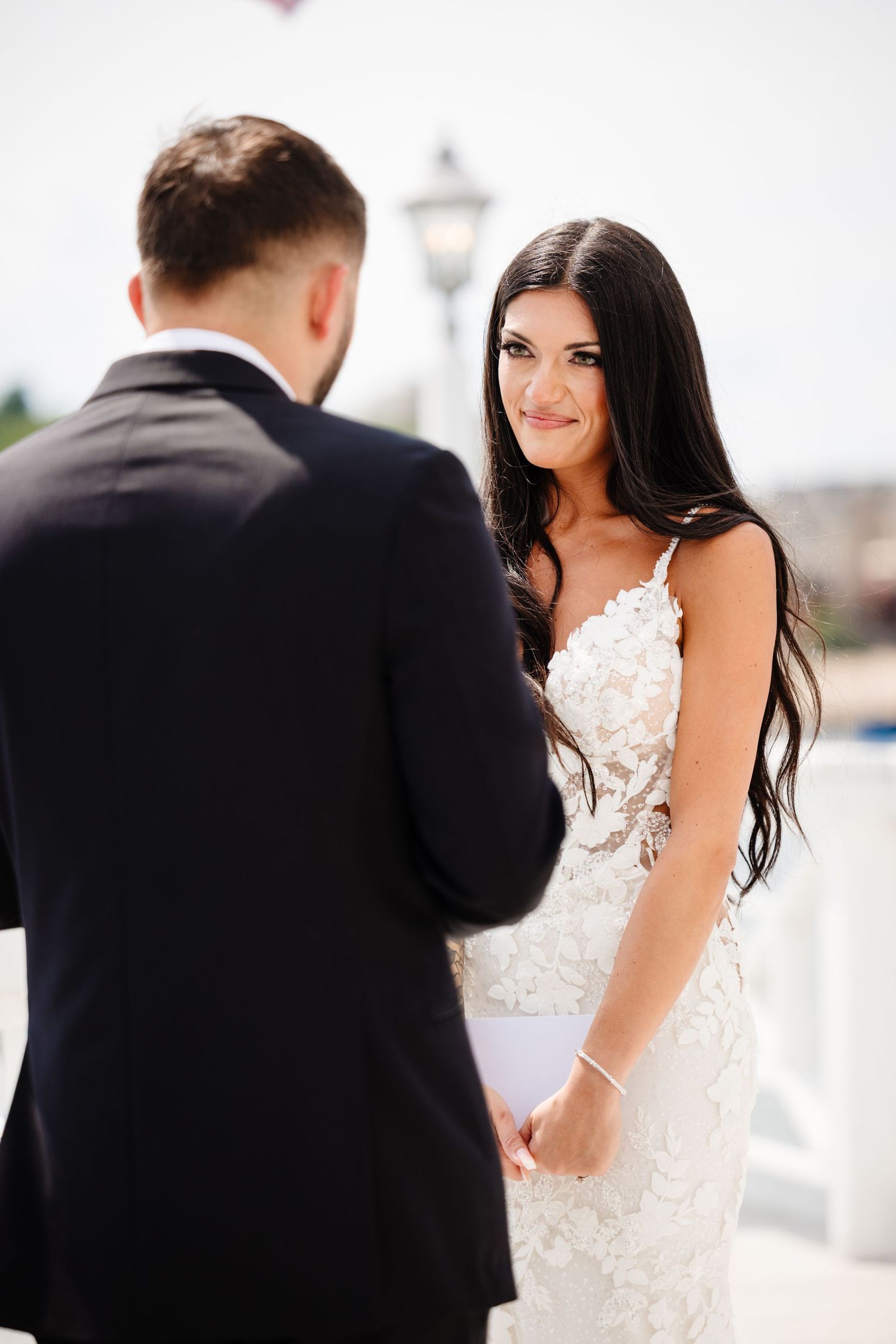 A bride in a lace wedding dress smiles at her groom during an outdoor wedding photoshoot.