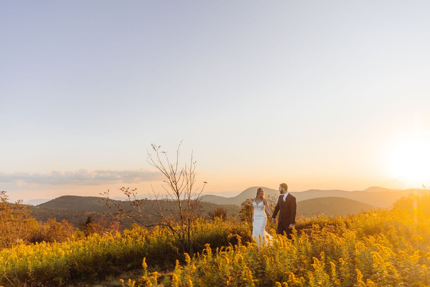 Wedding couple walks through golden meadow with mountain silhouettes at sunset.