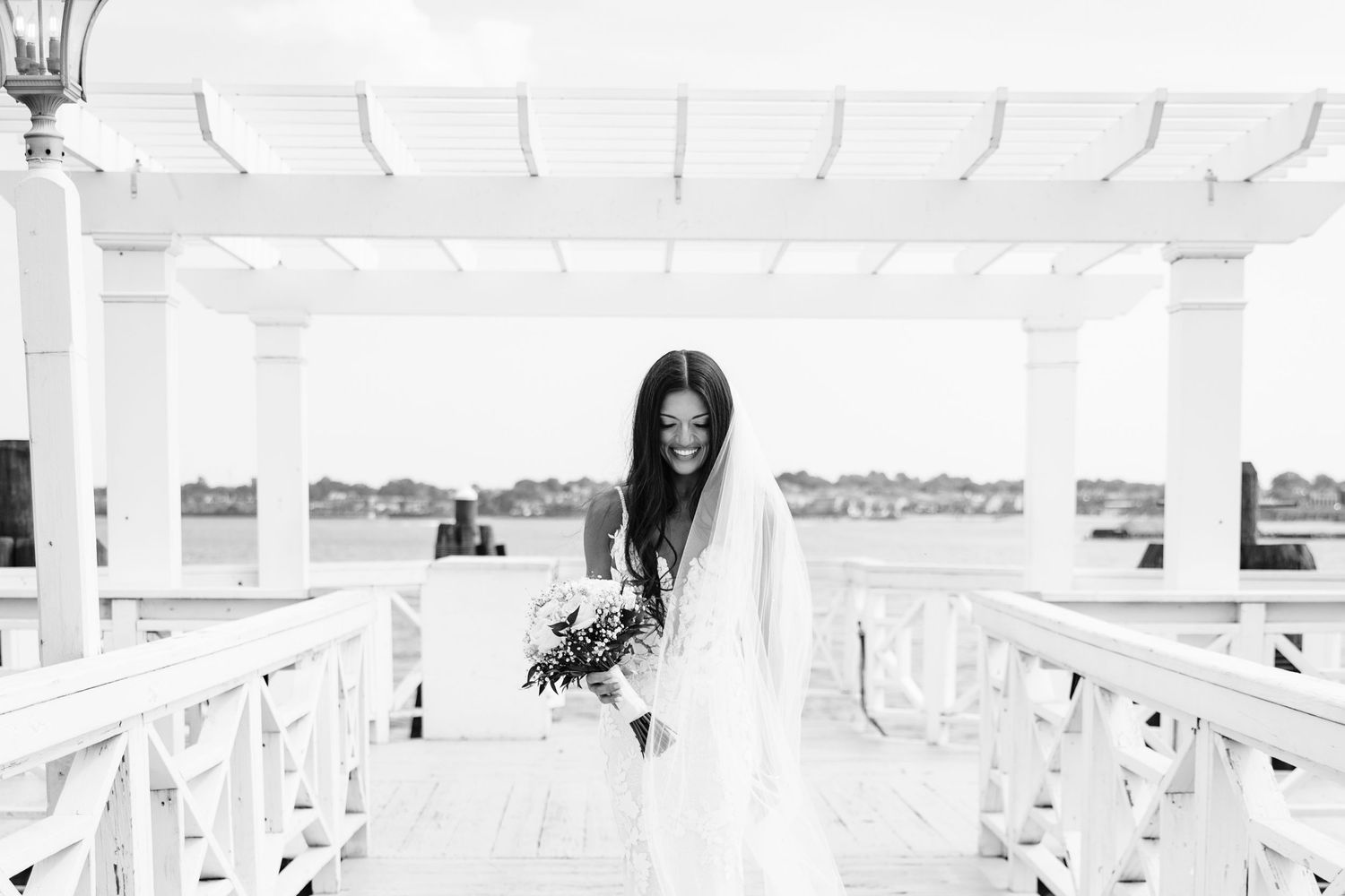Black and white image of bride holding bouquet while standing on white deck overlooking water.