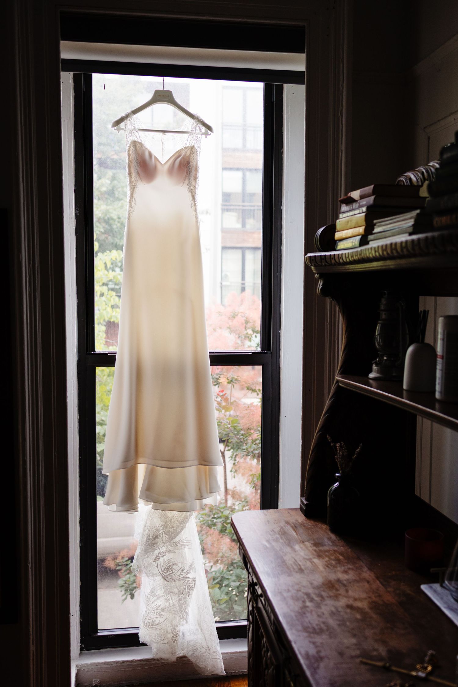 Elegant white wedding dress hanging in window frame with natural lighting.