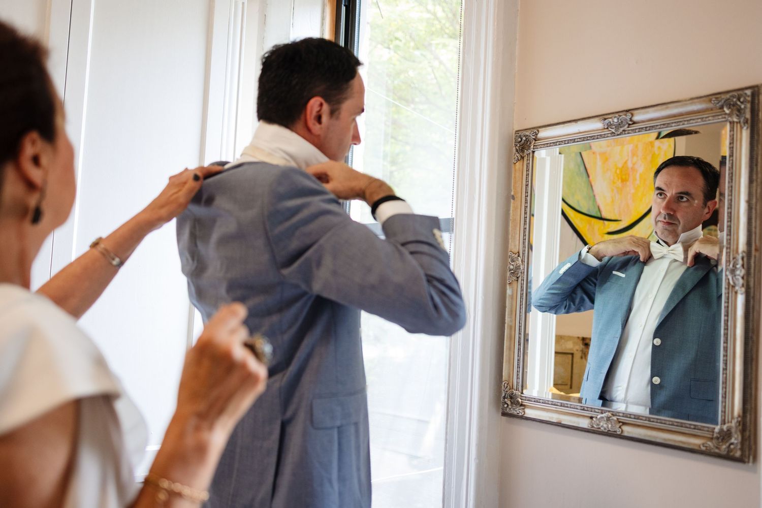Person looking in mirror while getting dressed in formal attire near window.