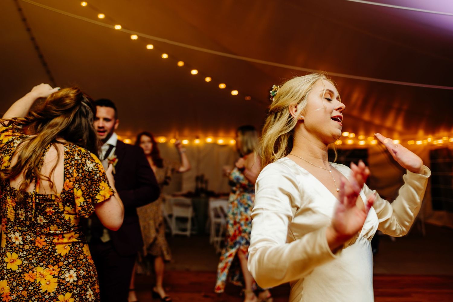 Wedding guests dance and celebrate under warm string lights at reception.