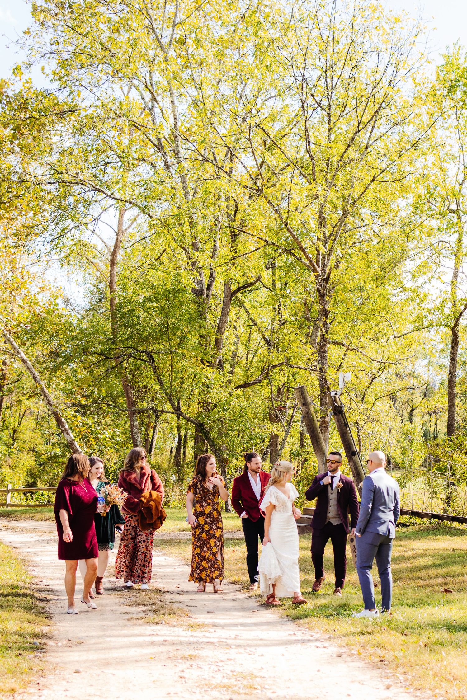Wedding party stands on a dirt path surrounded by golden autumn trees in a park setting.