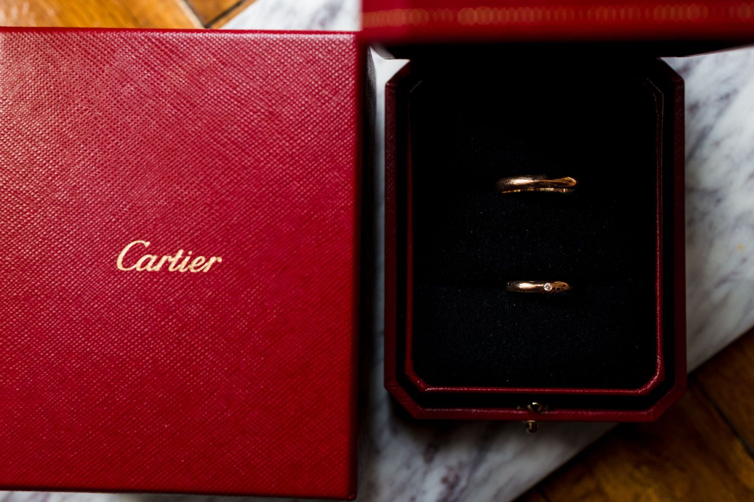 Two gold wedding rings displayed in red Cartier jewelry box on marble surface.