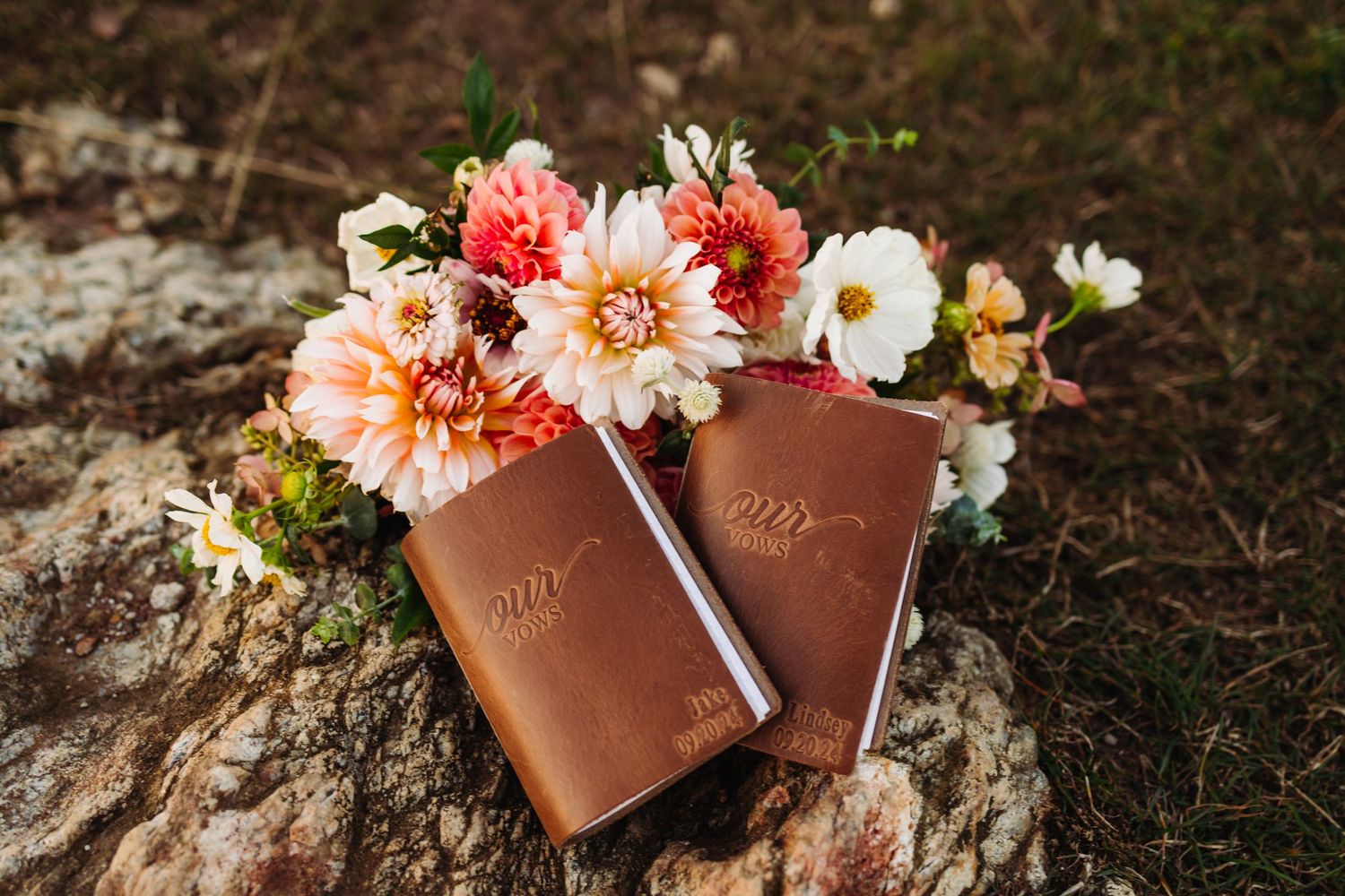 Two leather-bound vow books decorated with colorful dahlia and cosmos flowers on a rock.
