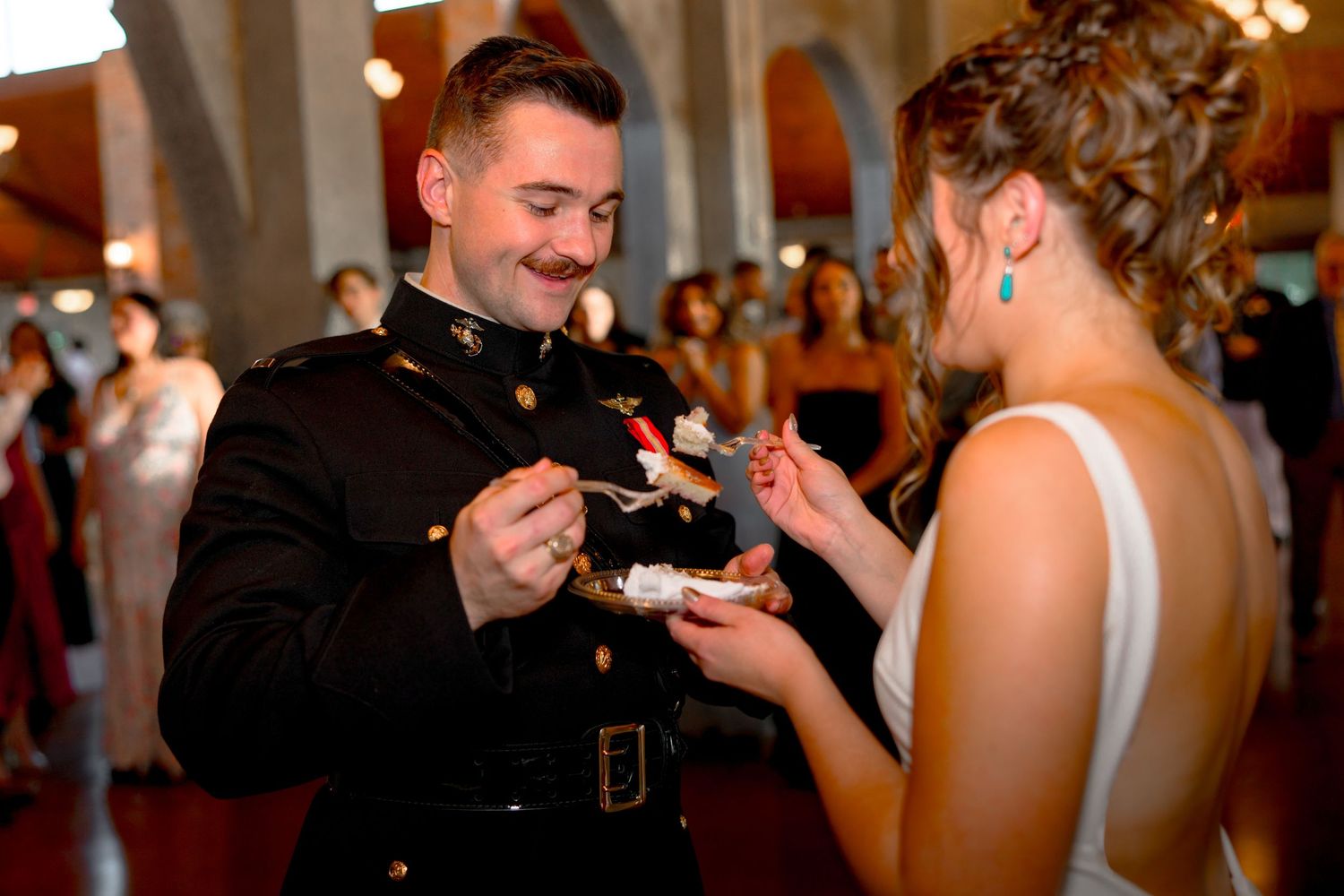 Groom feeding cake to his bride as they both smile on the dance floor. Their friends and family are behind them.