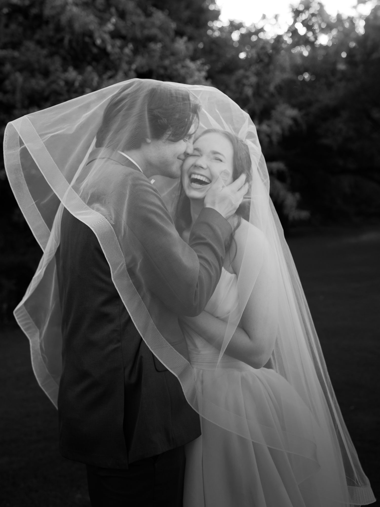 Bride and groom are smiling. He is kissing her cheek while they are under her veil photographed in black and white.