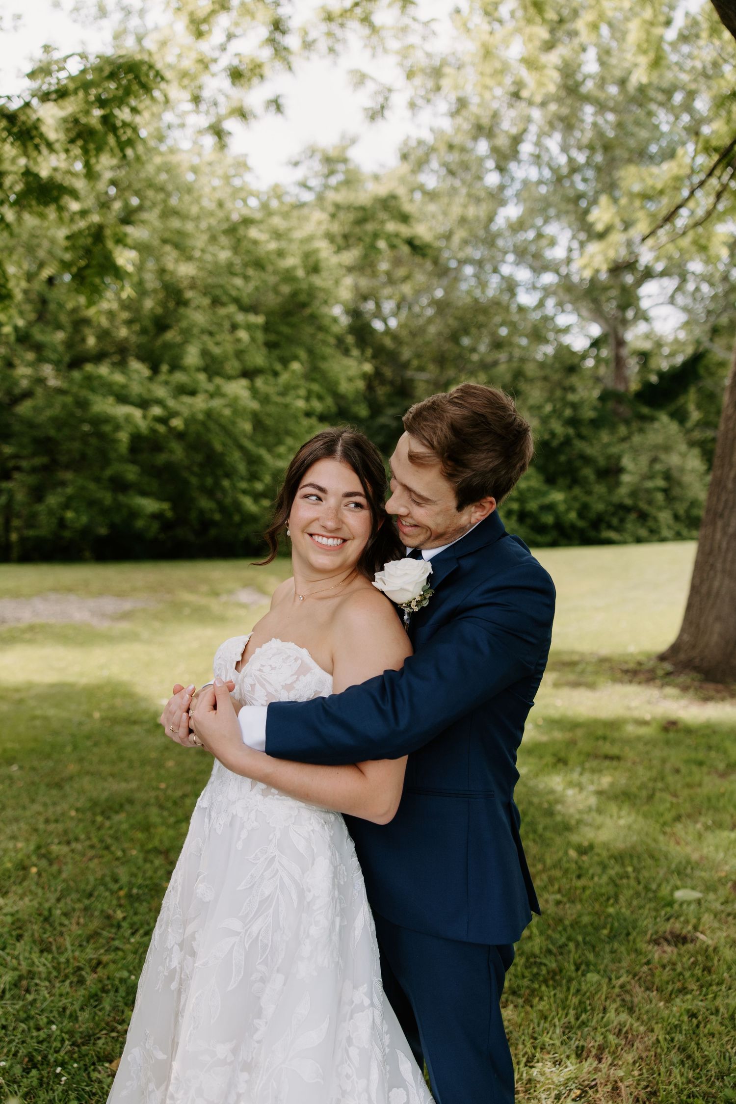 Bride and groom embracing outdoors in lush green park, bride in white lace gown smiling, groom in navy suit.