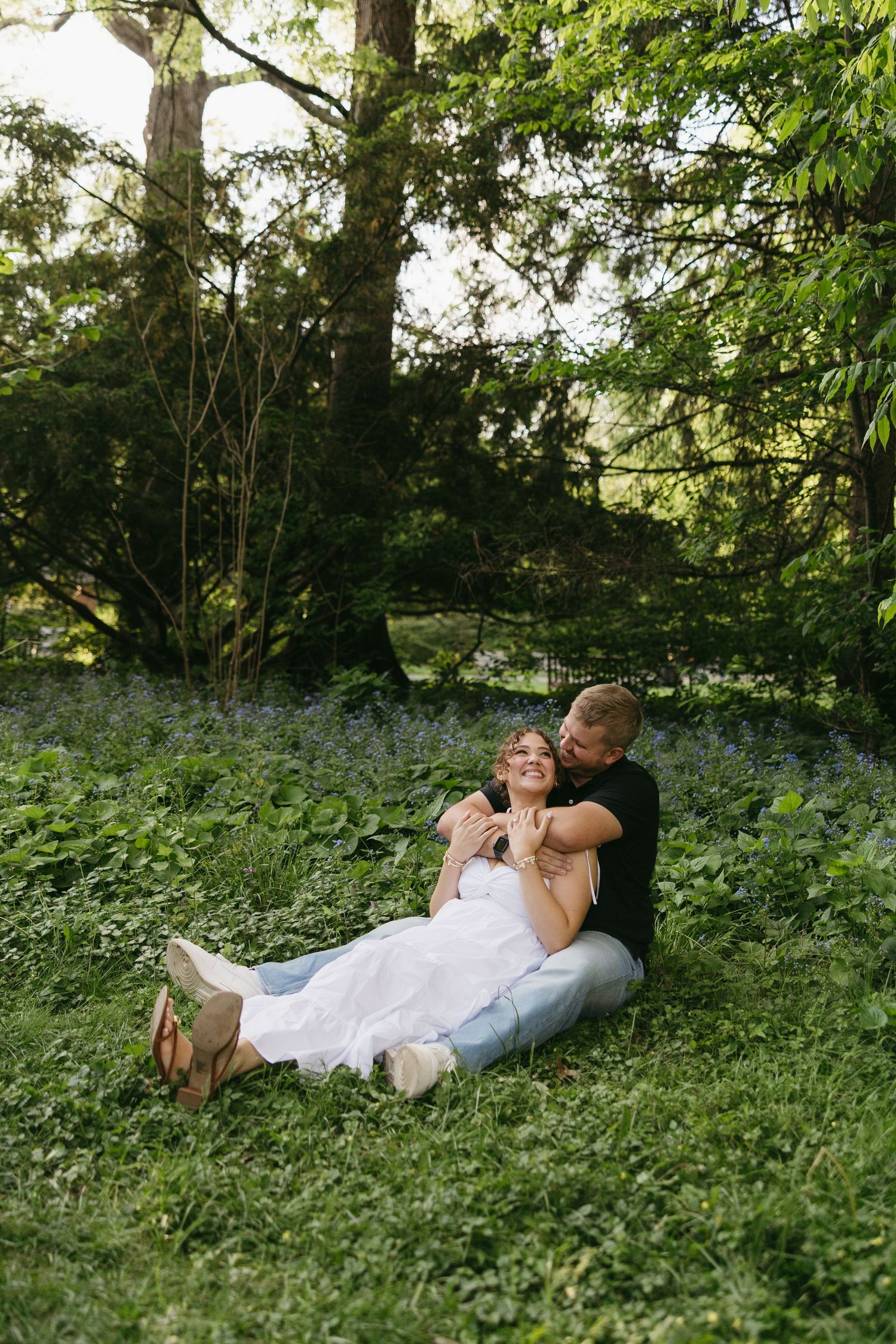 Couple sitting together in lush green forest, woman in white dress leaning against man in black shirt, surrounded by wildflowers.