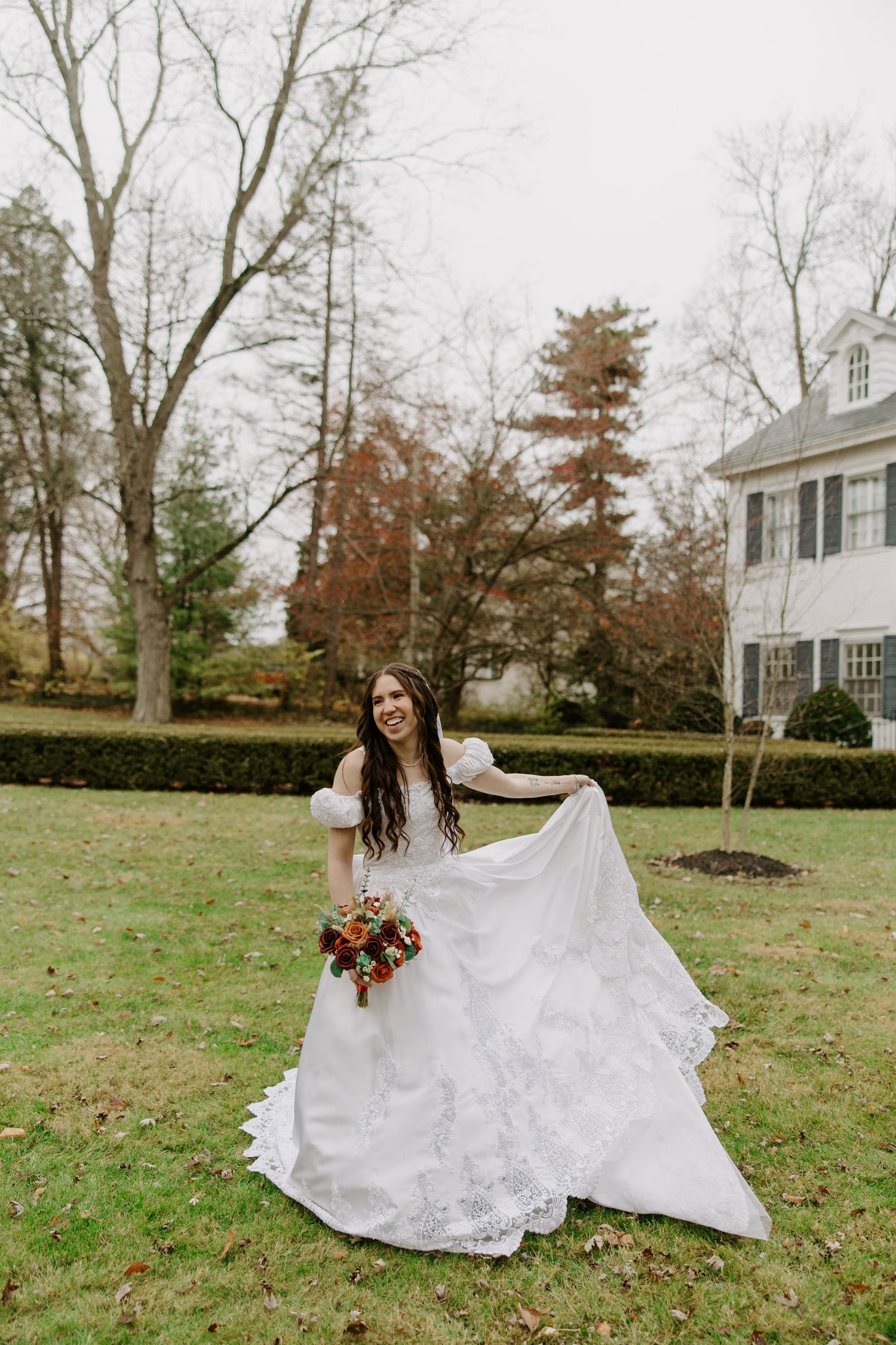 Joyful bride in white lace wedding gown twirls dress on green lawn with autumn trees and white house backdrop.