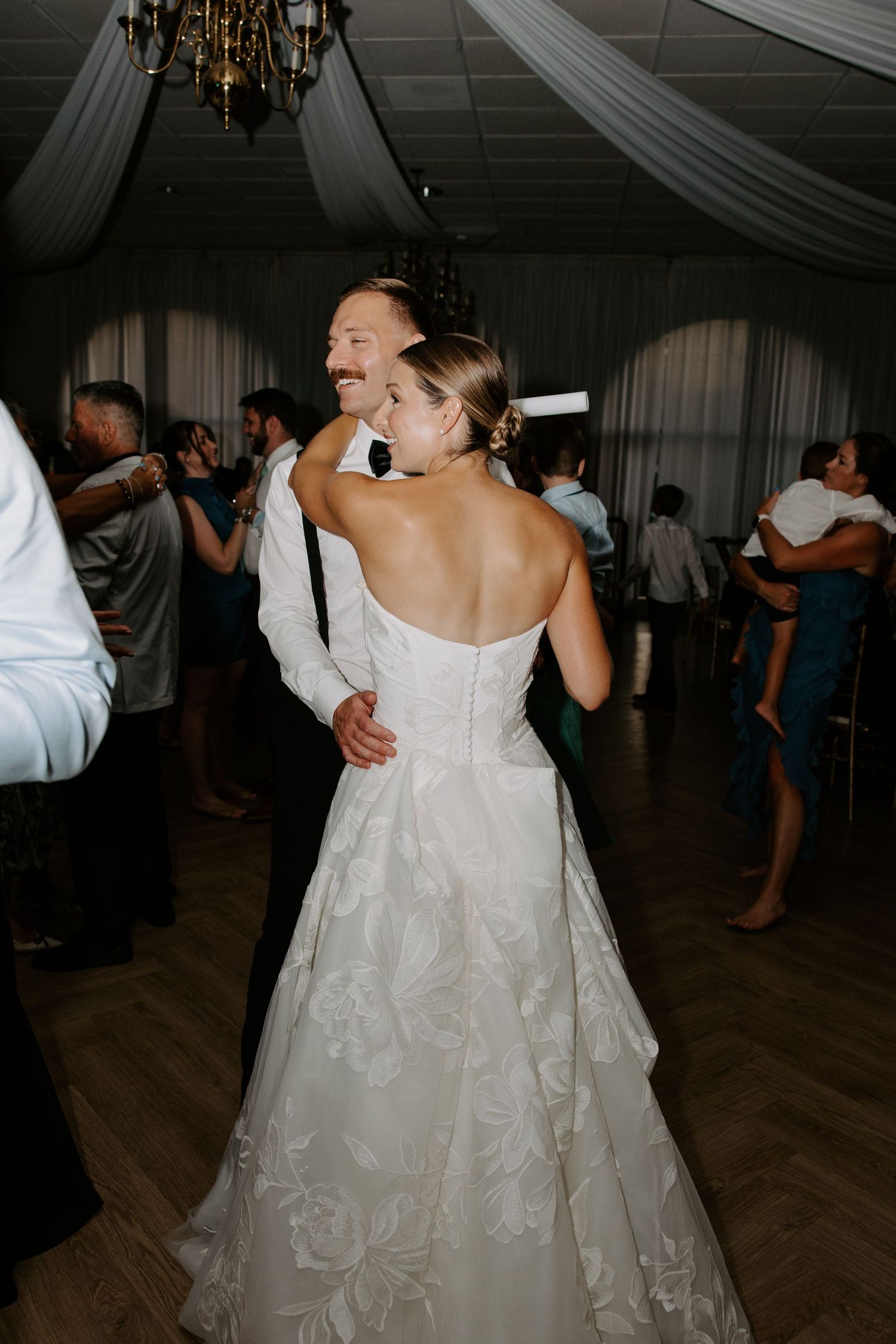 Bride in strapless lace gown dancing with guest at elegant wedding reception with draped ceiling and chandelier.