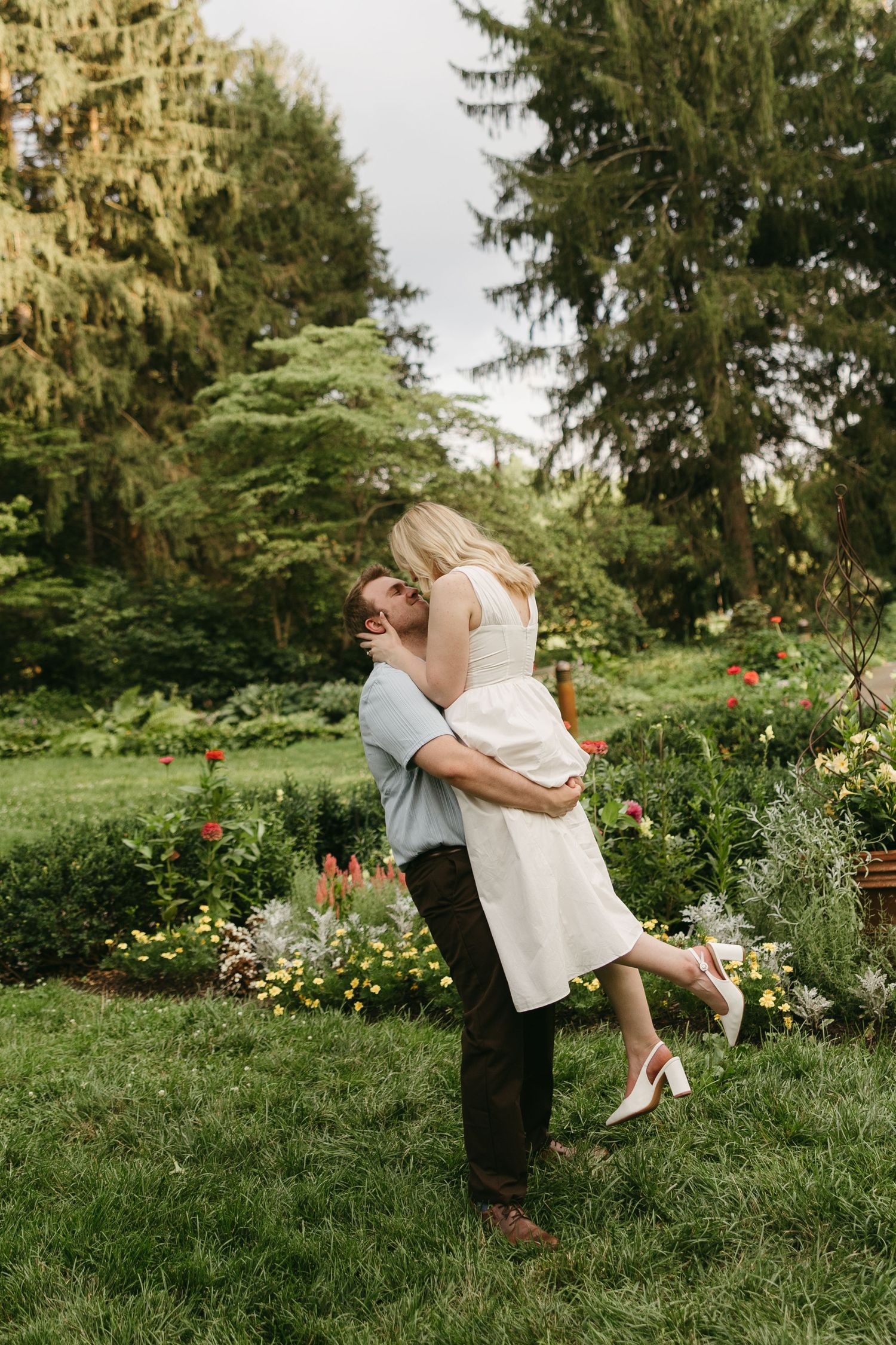 Couple sharing a romantic lift embrace in a lush garden with colorful flowers and tall evergreen trees.