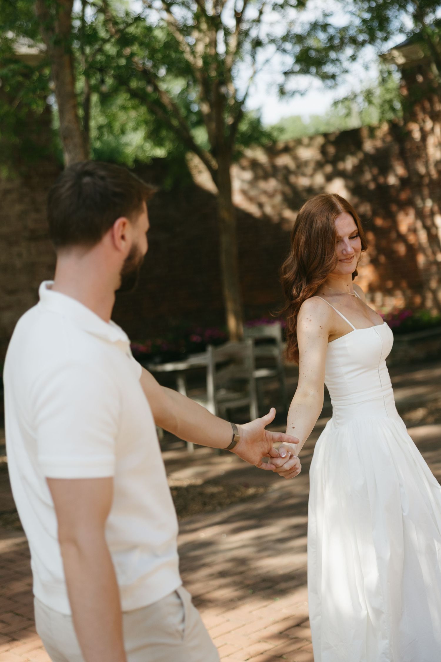 Bride in white spaghetti-strap gown leads groom by the hand in a shaded garden.